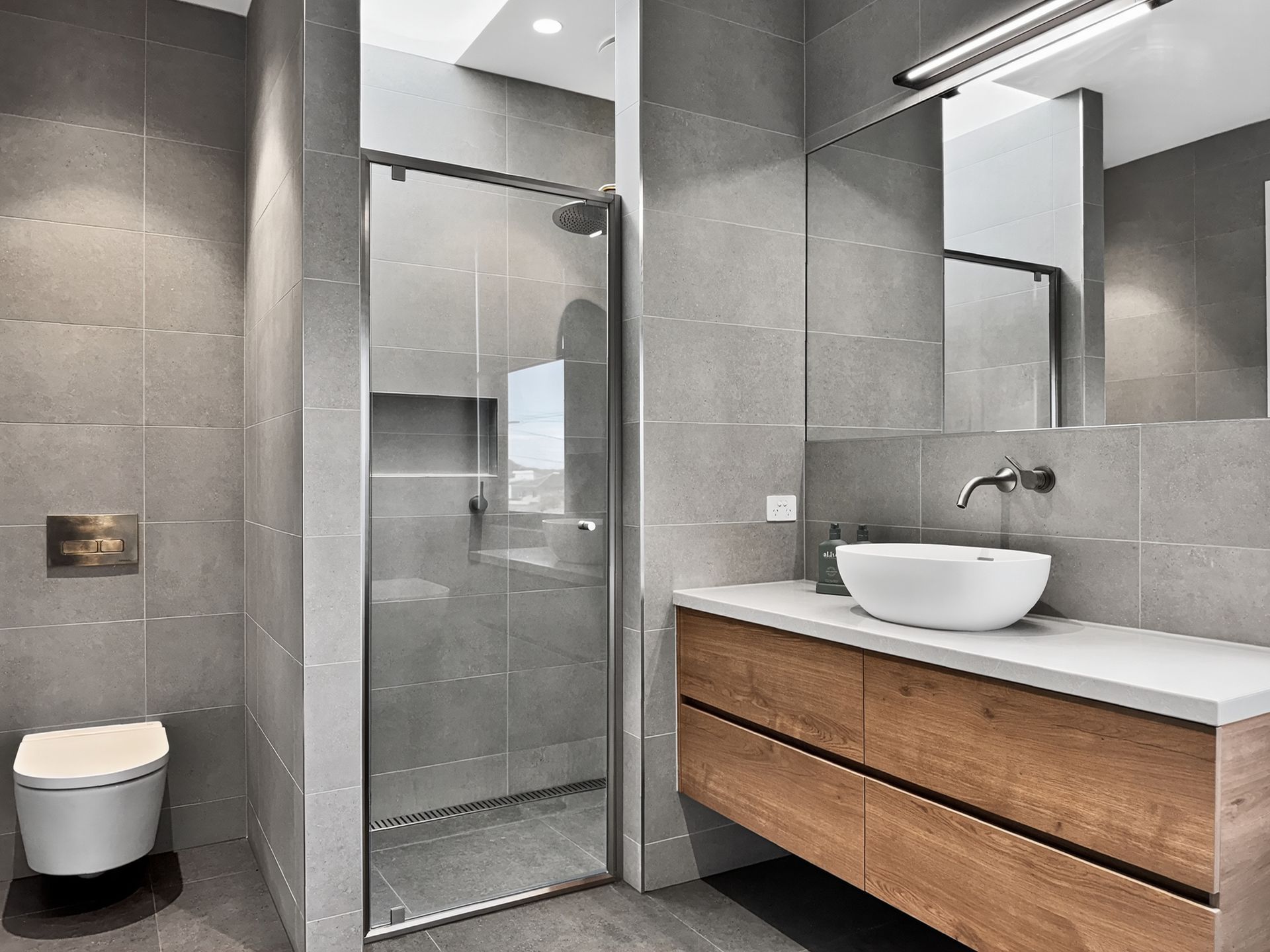 Modern bathroom with gray tiles, glass shower, wooden vanity, and a white sink.