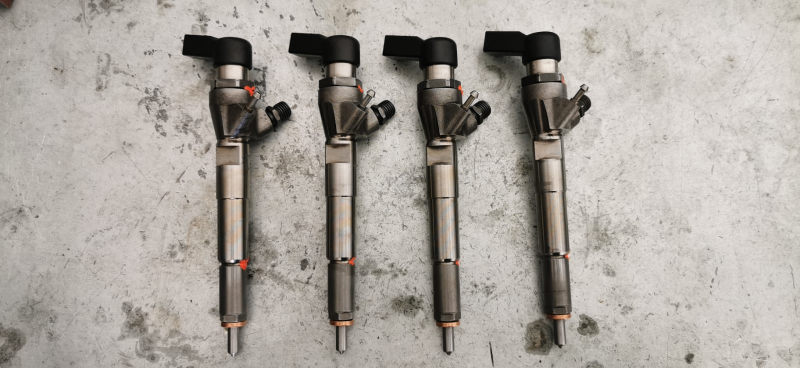 Quattro iniettori di carburante disposti su una superficie grigia. Sono cilindrici, argentati, con la parte superiore nera.