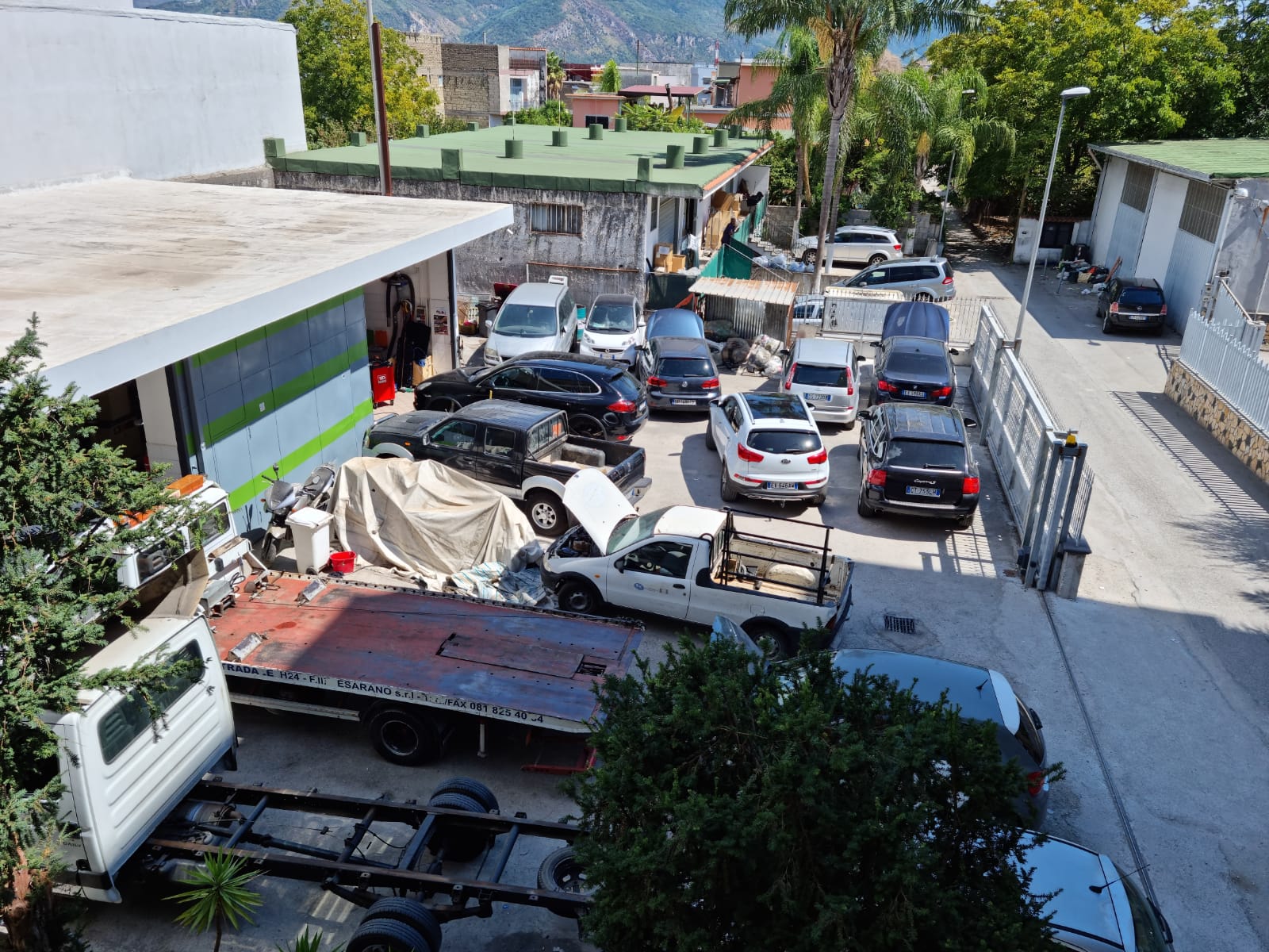 Auto parcheggiate in un'officina all'aperto; diversi veicoli sono in riparazione, con un camion con pianale di fronte.