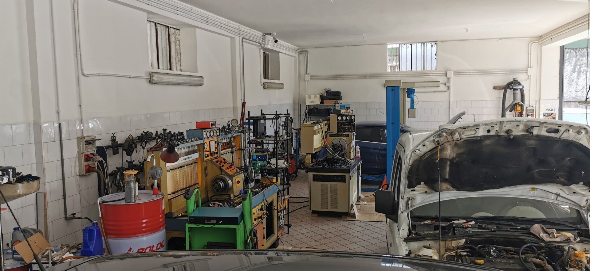 Un'officina con pezzi di ricambio, macchinari e un'auto con il cofano aperto. Pareti bianche, illuminazione dall'alto.