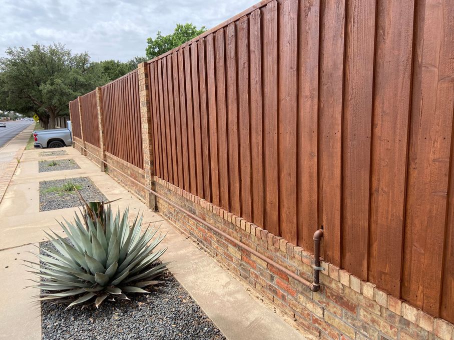 Wooden fence with brick base, next to a sidewalk and a large succulent plant.