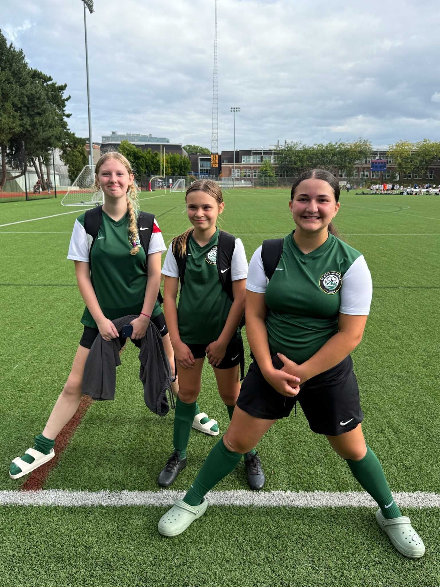 Three people in green and black athletic attire pose on a soccer field, smiling.