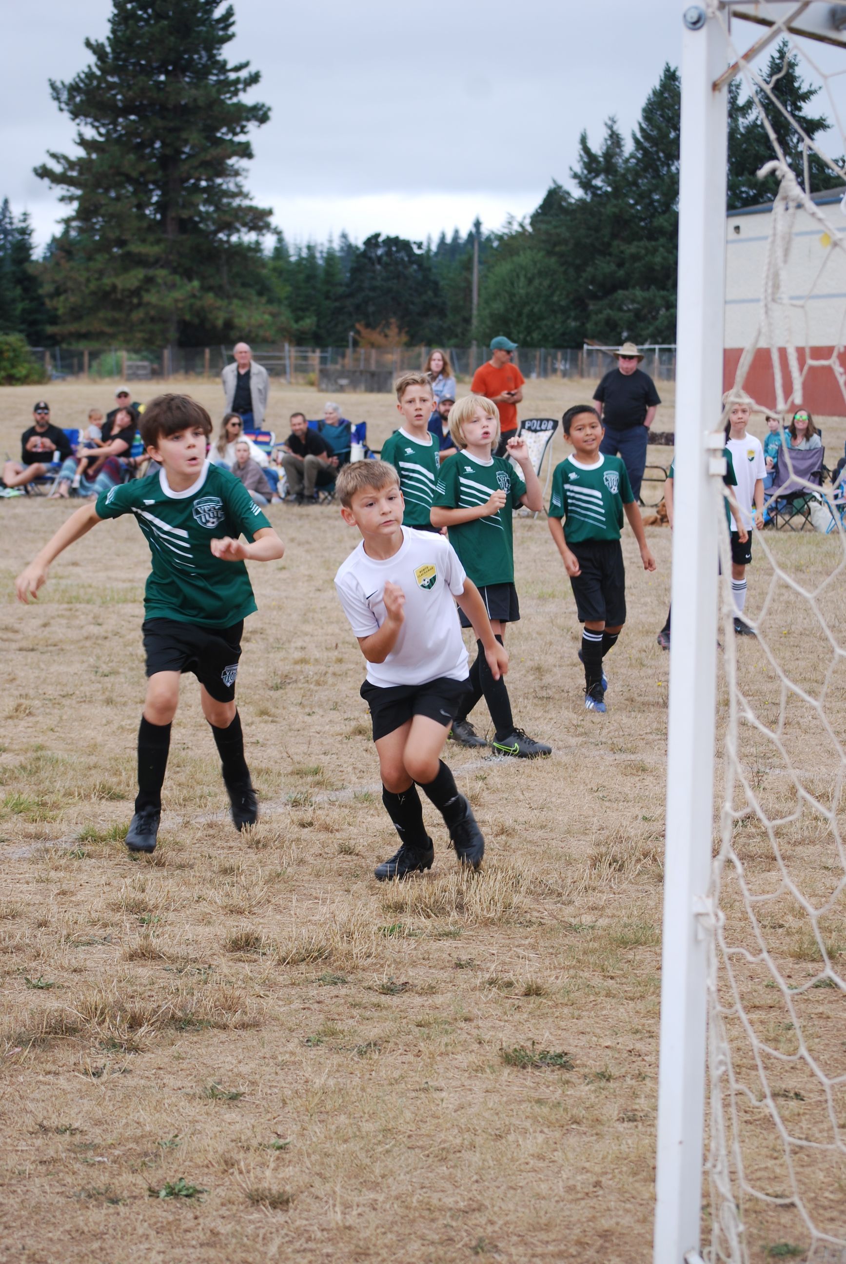 Kids in soccer uniforms running toward a goal on a field, with spectators in the background.