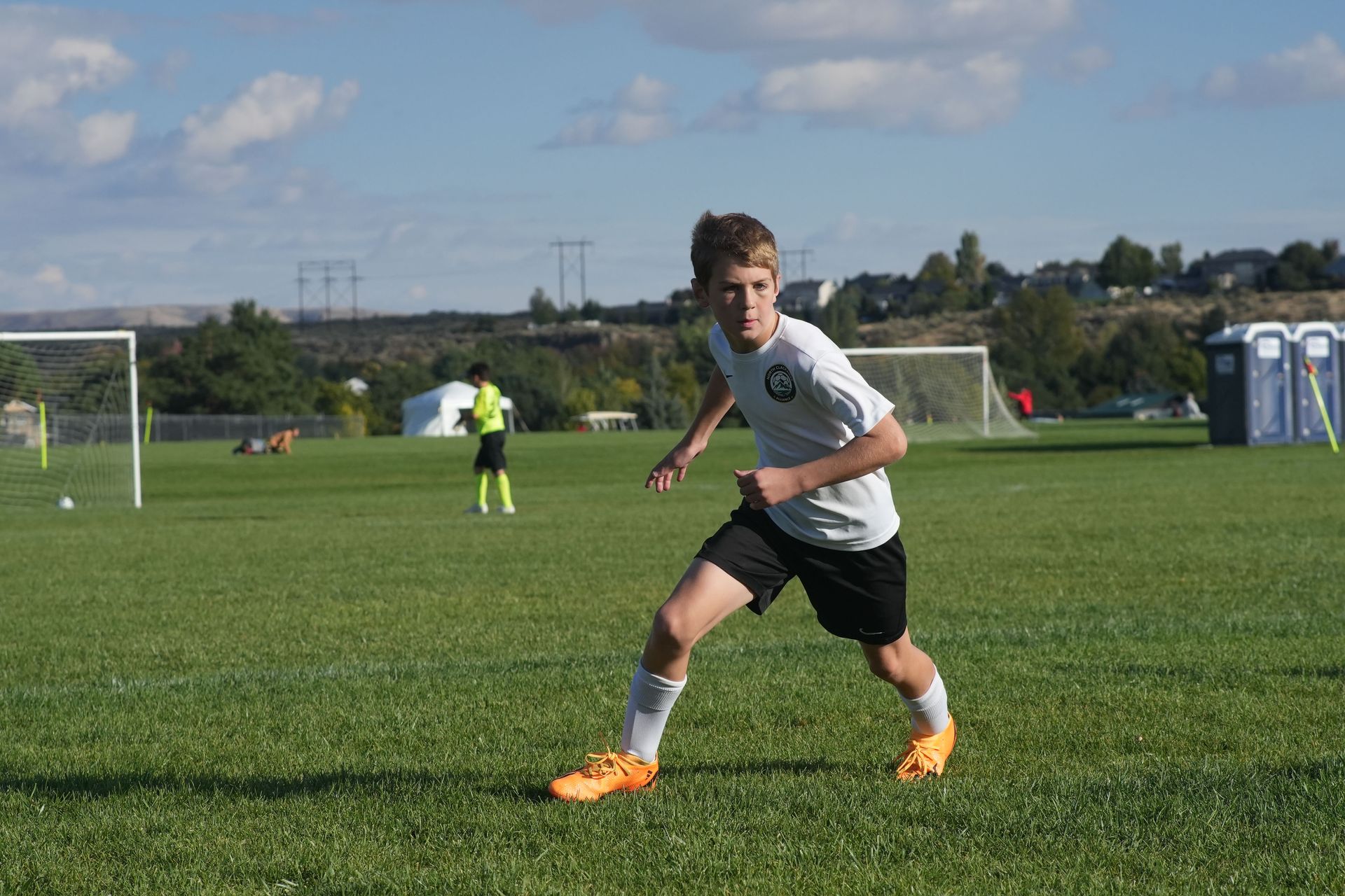 Boy in soccer uniform running on a green field towards the right. Other players and goals in the background. Sunny day.