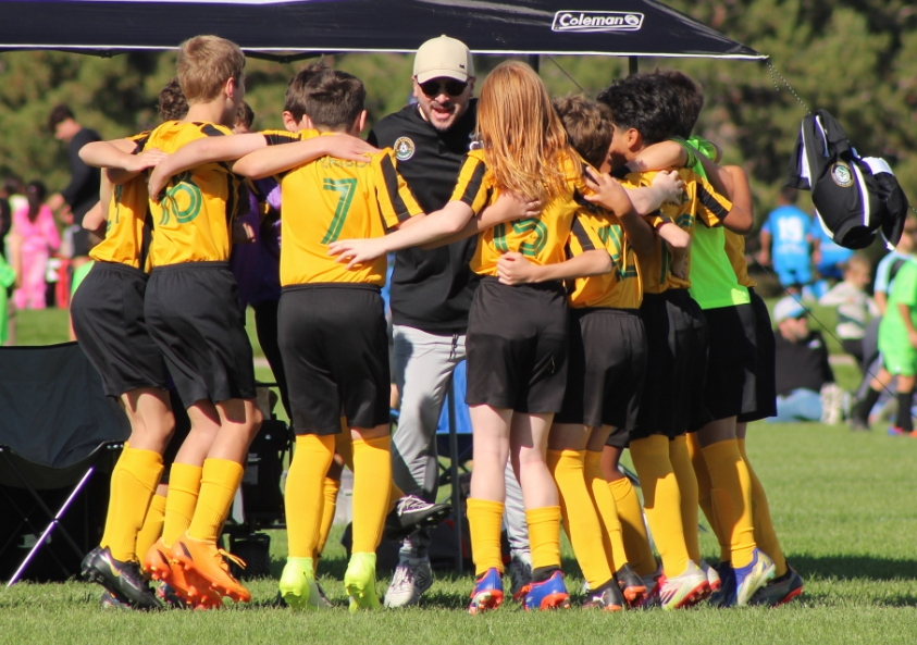 Soccer team in yellow and black uniforms huddling with their coach on a green field.