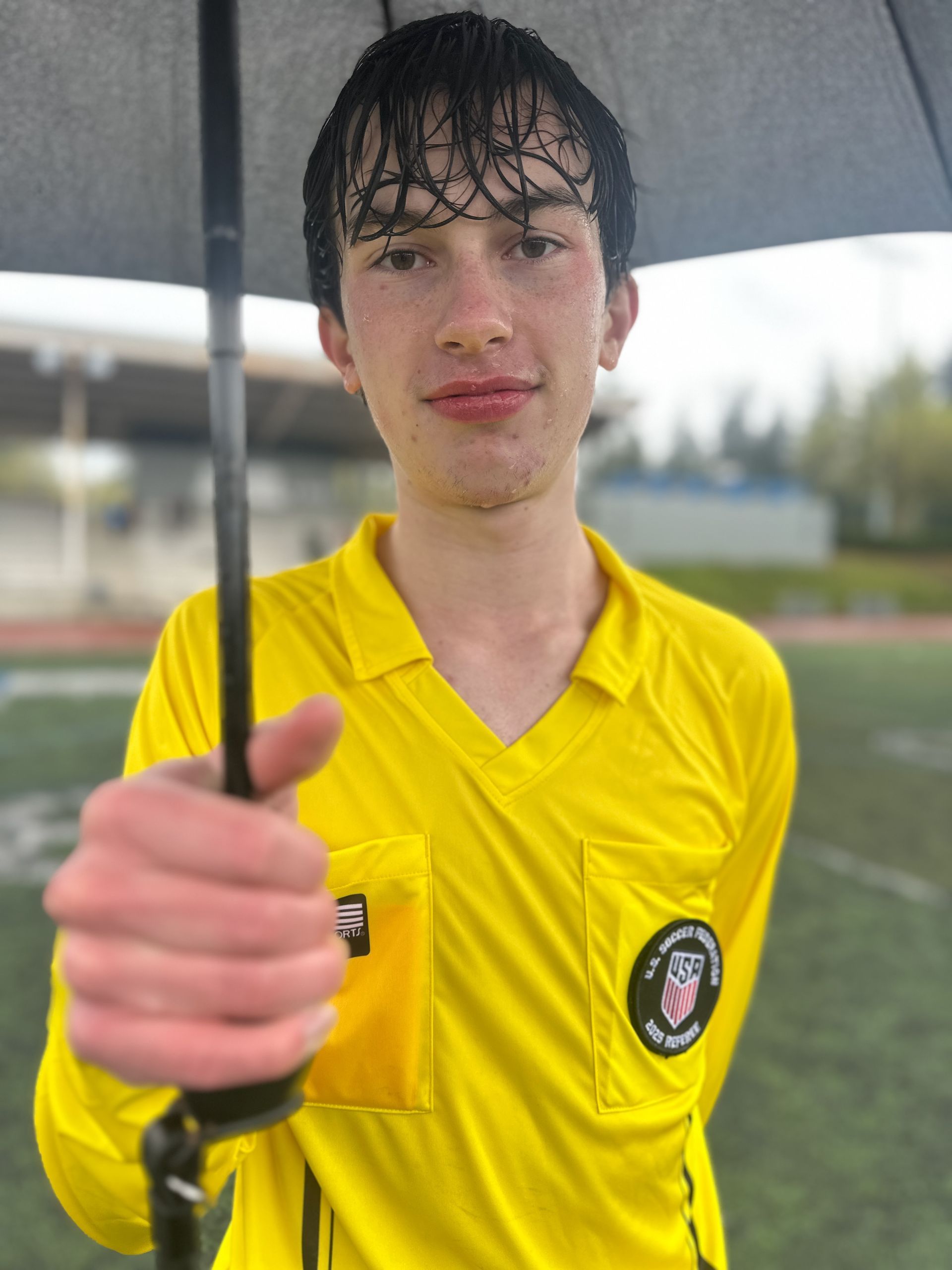 Soccer referee holding an umbrella, wearing a yellow uniform, smiling slightly outdoors.