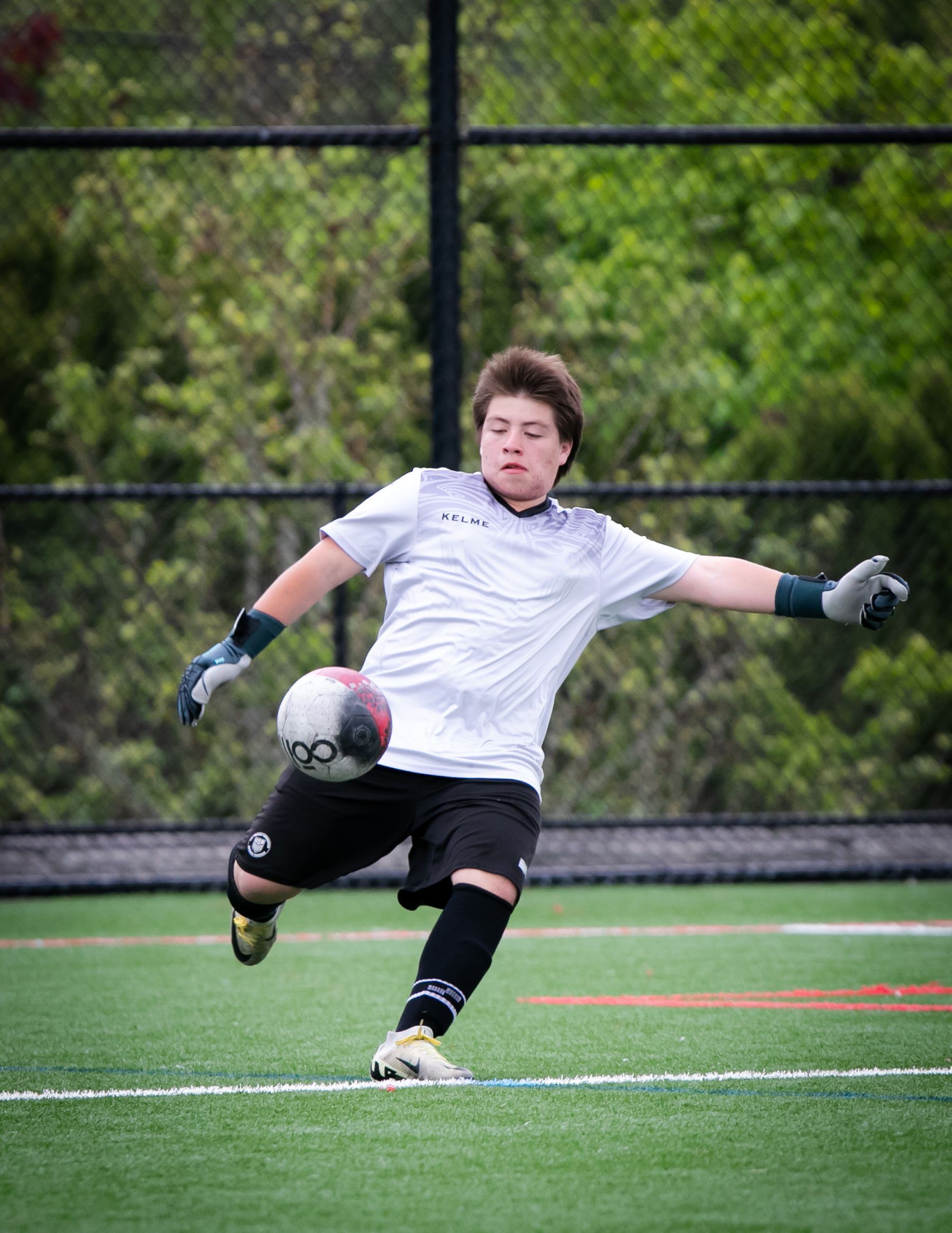 Soccer goalie kicks ball on a green field. Wearing gloves, white jersey, black shorts, and socks.