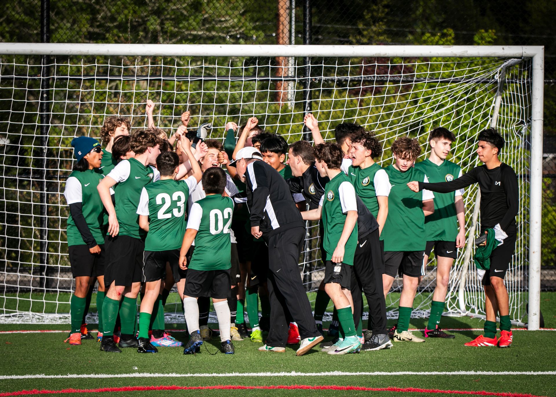 Soccer team celebrating victory in front of the goal, green and black uniforms, arms raised, on a green field.