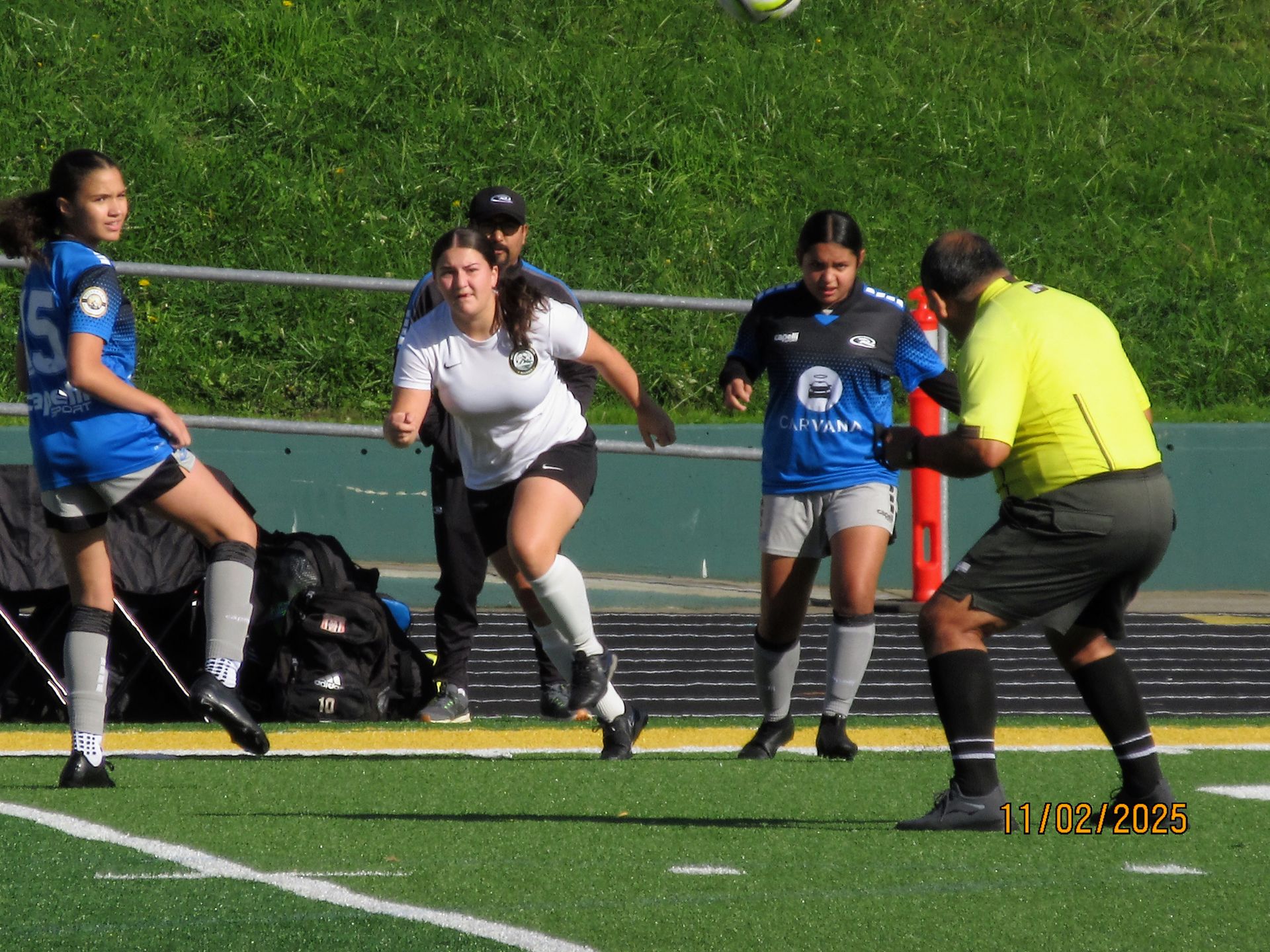 Soccer players on a field with a referee. One player in white runs towards the ball.