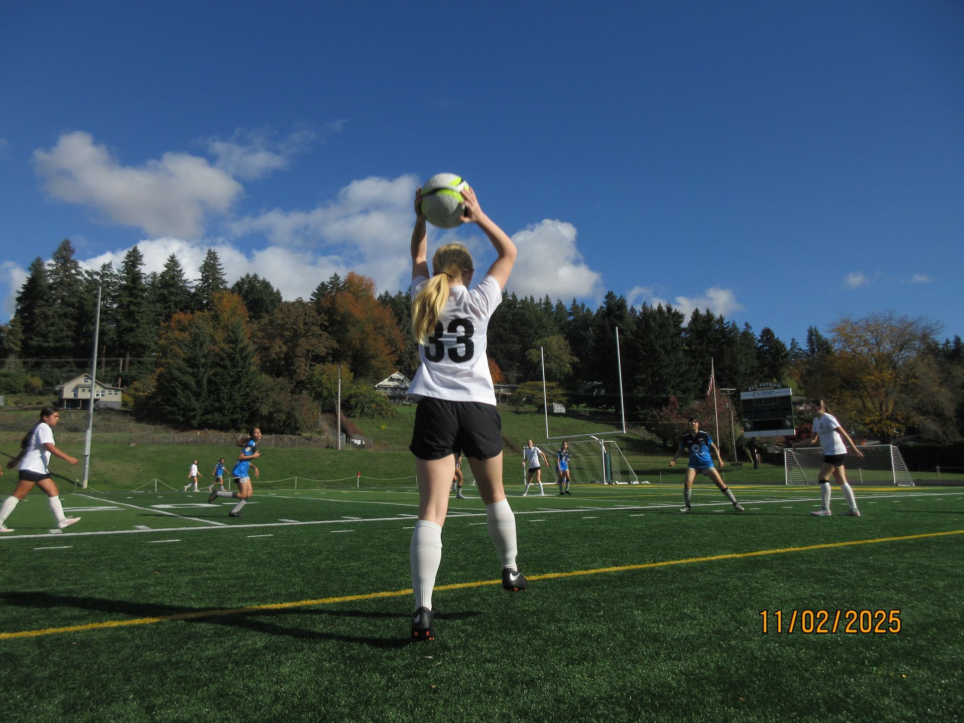 Soccer player in white jersey with number 33 throws the ball. Other players and trees in the background on a field.