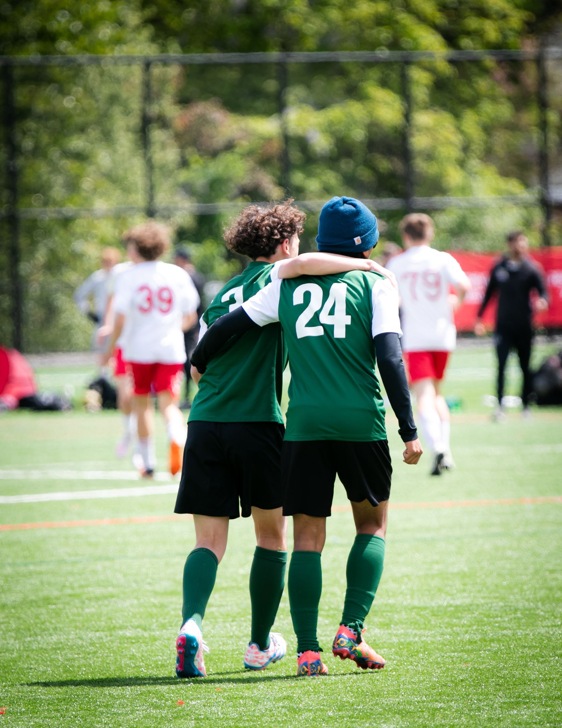 Two soccer players in green jerseys embrace on a green field. Players in red jerseys are in the background.