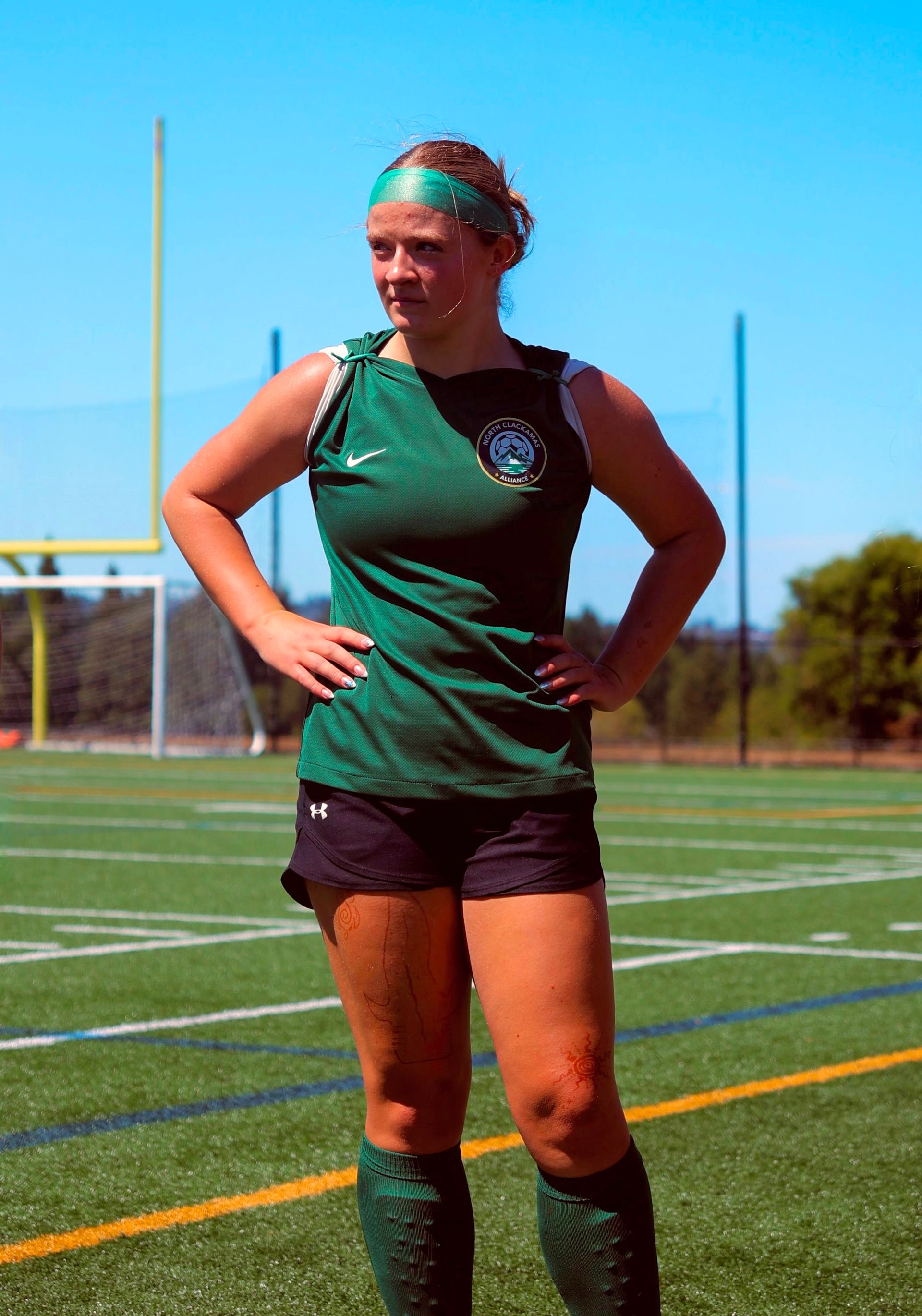 Woman in green athletic wear on a turf field, hands on hips, looking to the side.