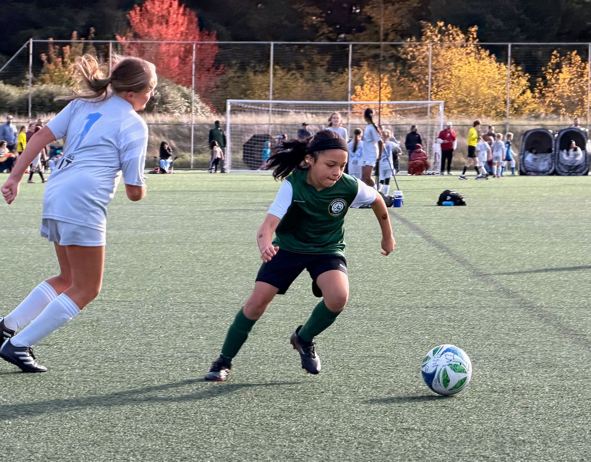 Soccer players on green field, one in green jersey dribbling ball, opponent in white, referee in yellow behind.