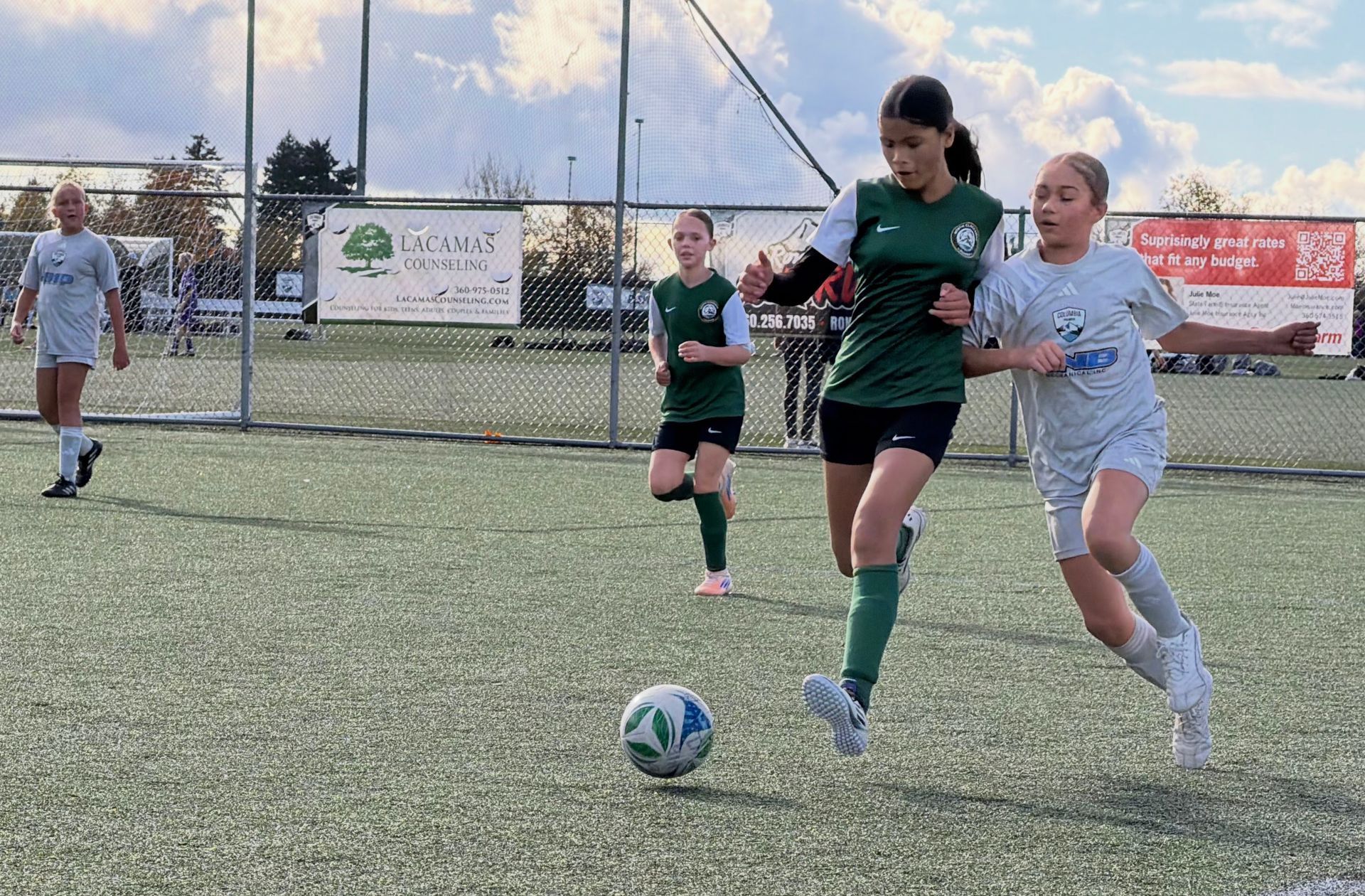 Soccer players on a field: one in green dribbles the ball, another in grey attempts to block. Other players in the background.
