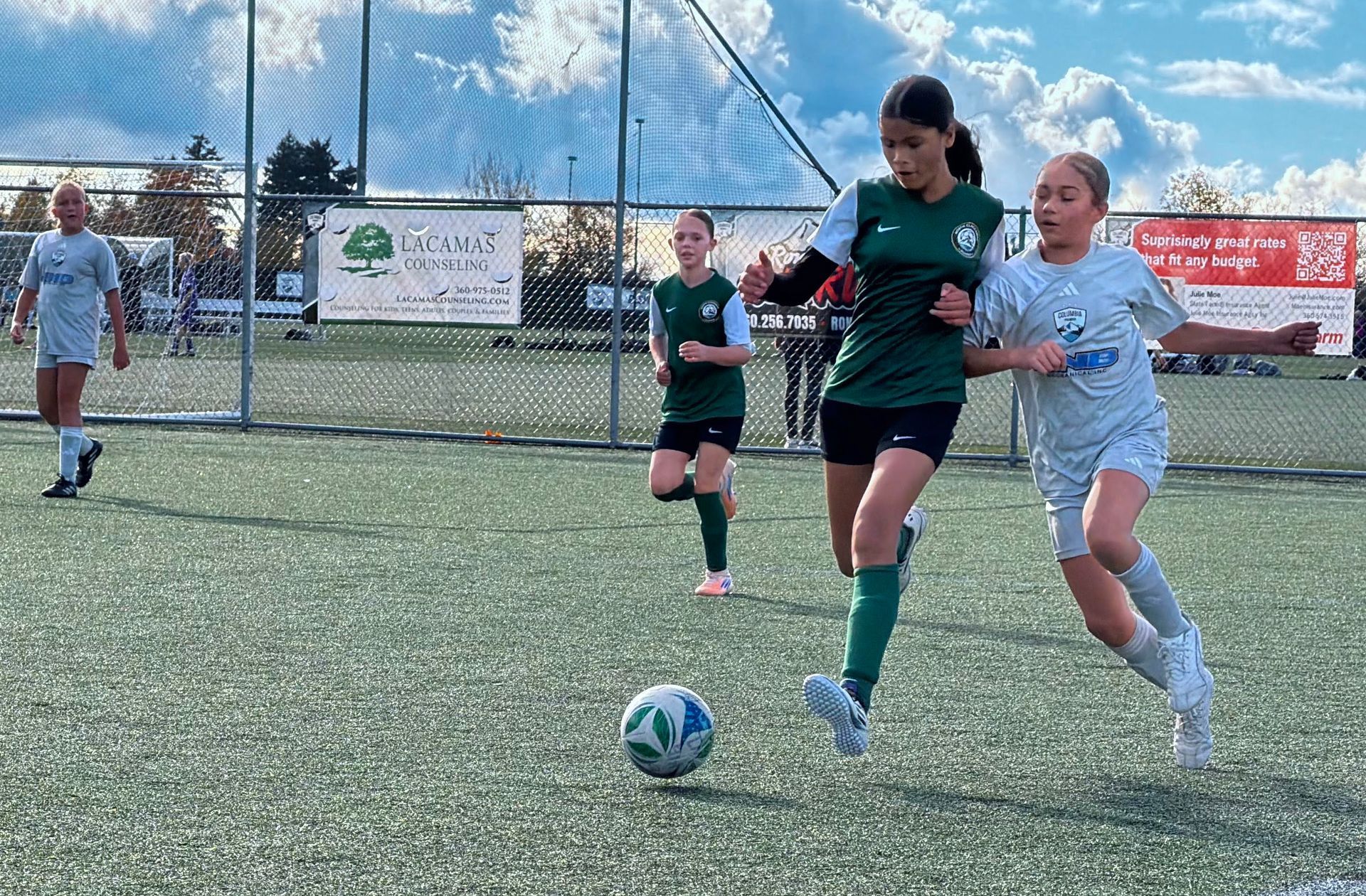 Soccer players on a green field. A player in green dribbles past another in gray. Cloudy sky.