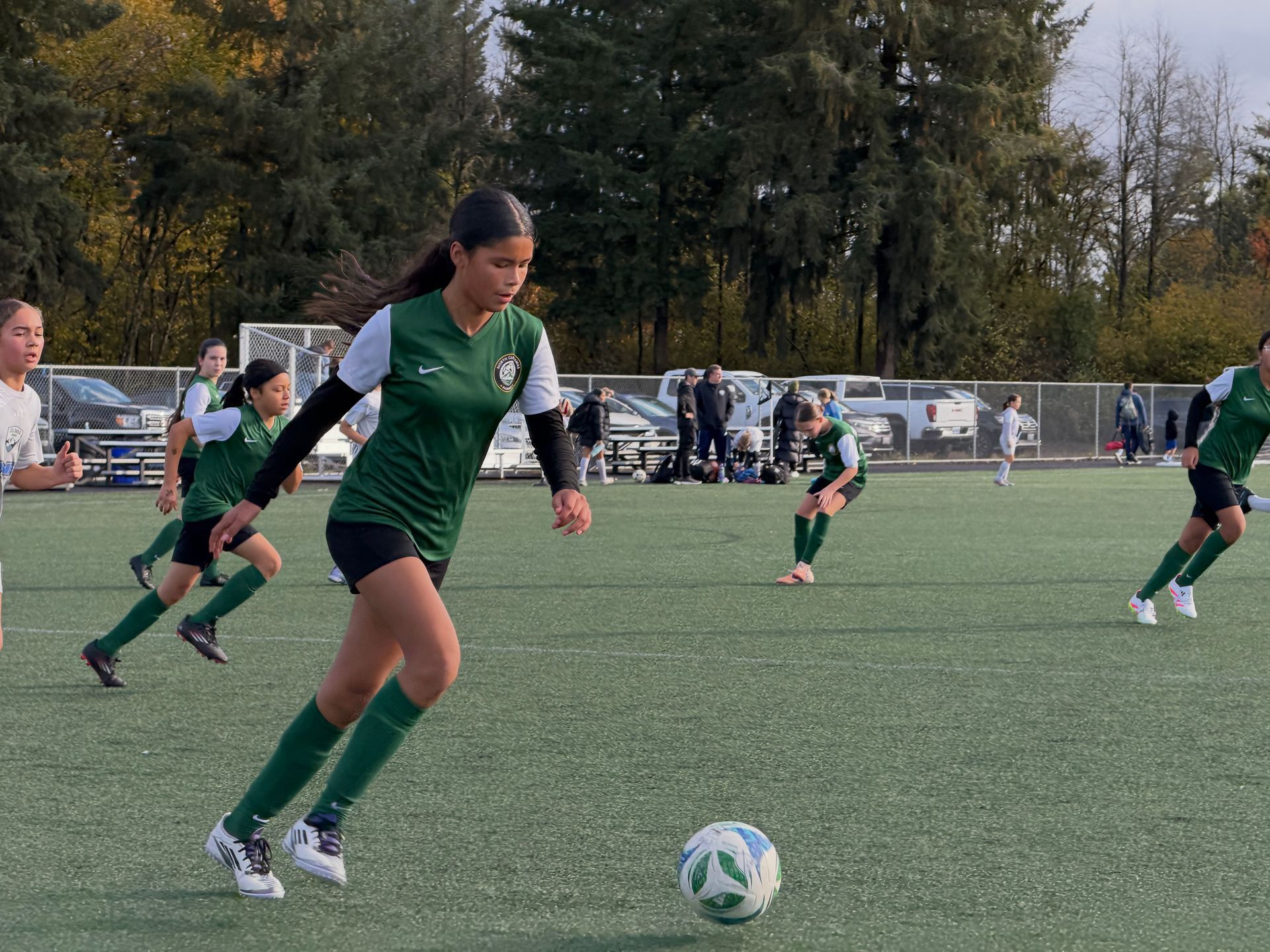 Soccer player in green jersey dribbles the ball on a green field. Other players watch in background.