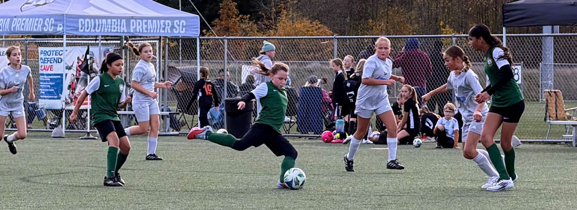 Soccer players on a field; one in green kicking the ball. Others in light uniforms run nearby.