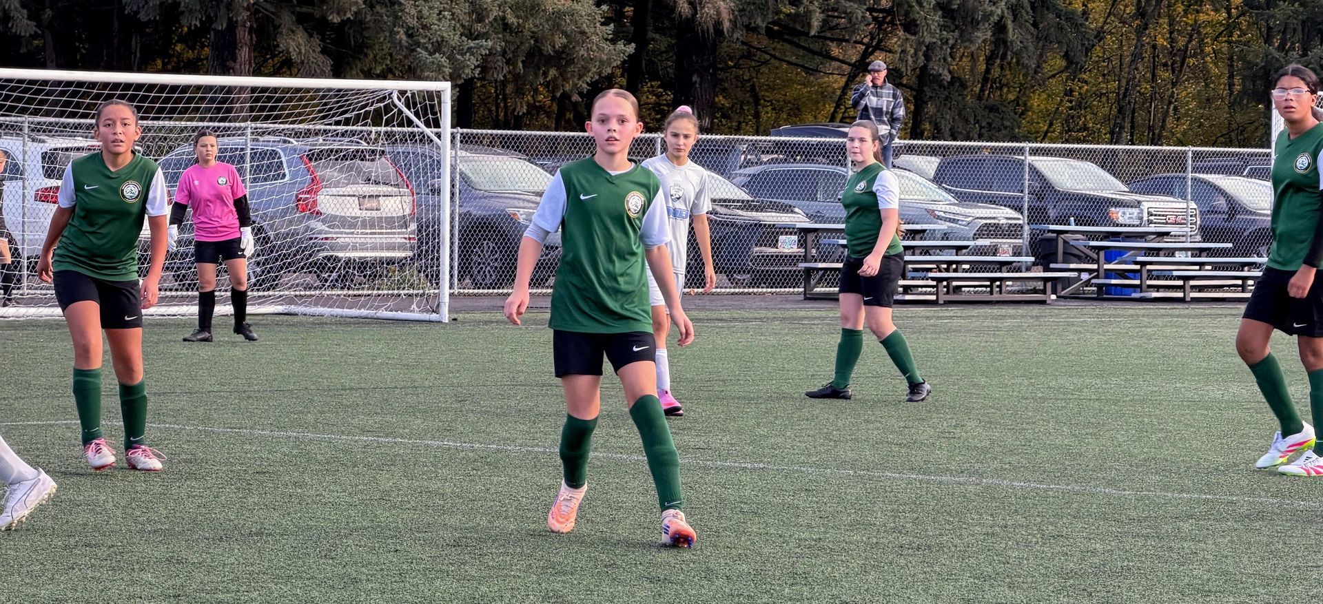 Soccer players on a green field. Players in green and white uniforms are facing towards the camera. The goal is in the background.