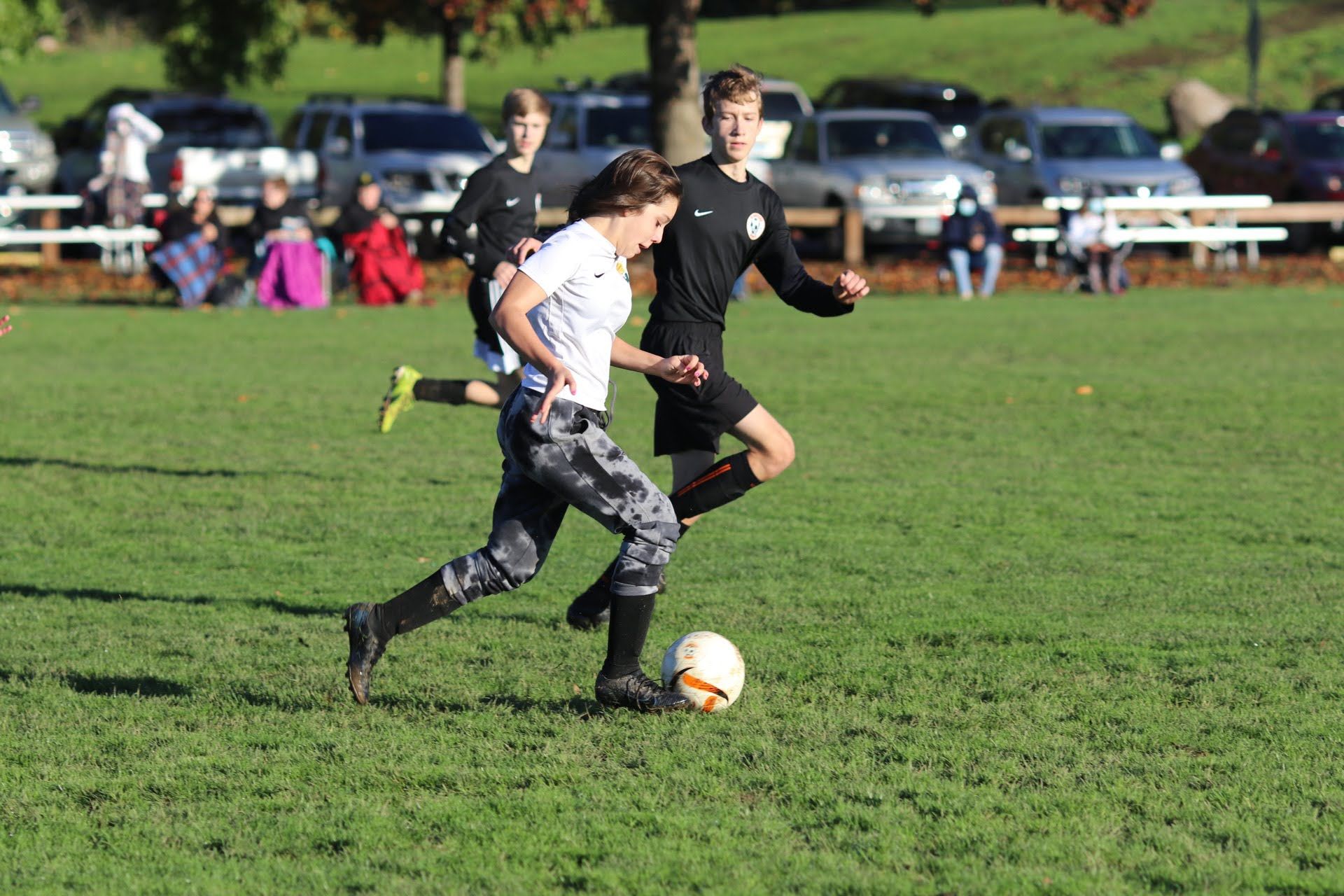 Soccer players on a grassy field. One in white shirt dribbles the ball, another in black defends. Sunny day.
