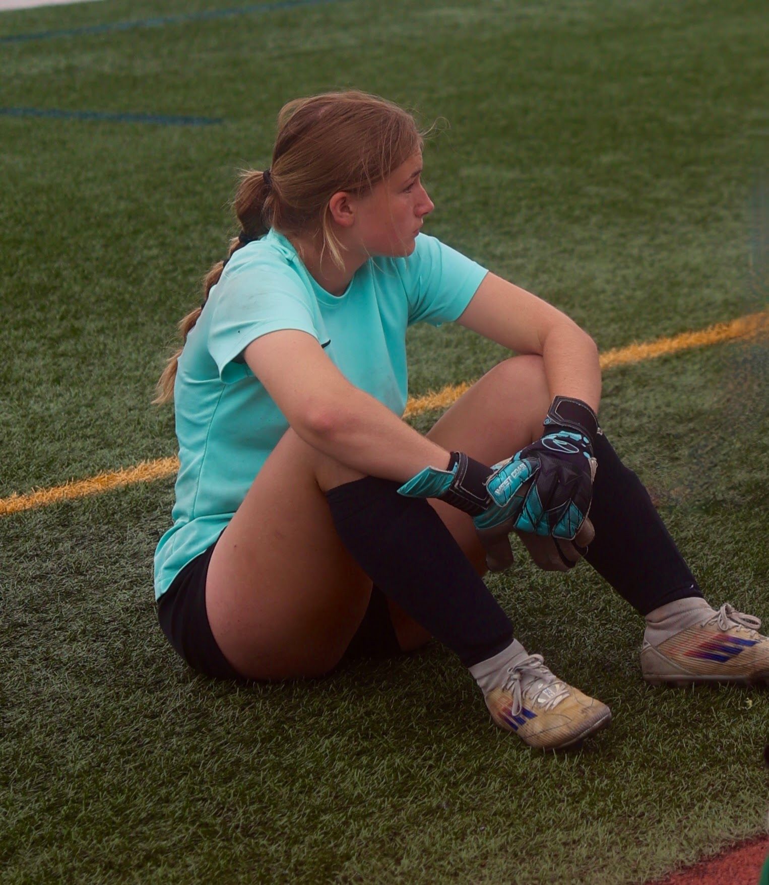 Soccer player in a teal shirt sits on the field, wearing gloves and socks, looking to the side.
