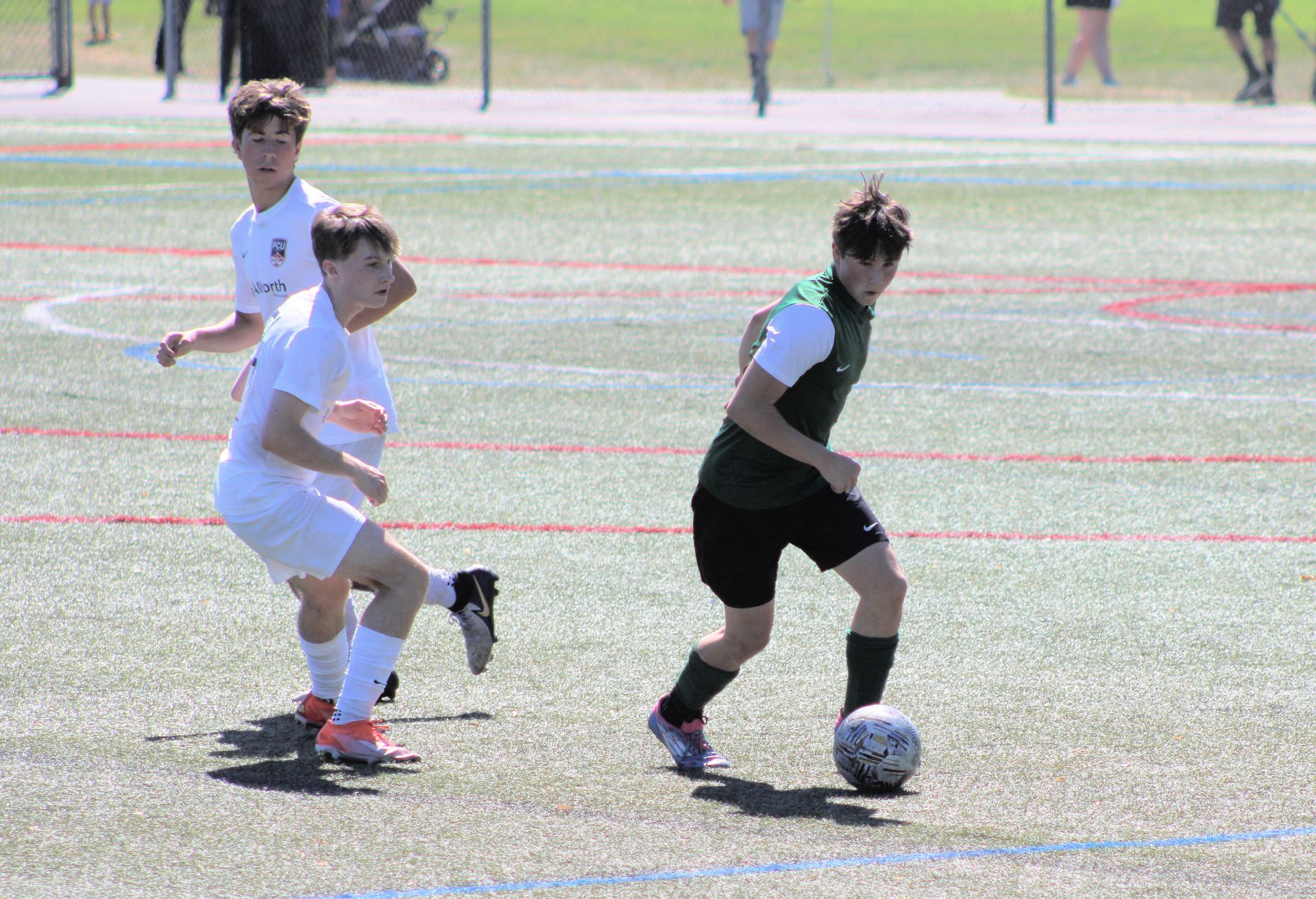 Soccer players on field: one in green dribbles ball, two in white nearby.