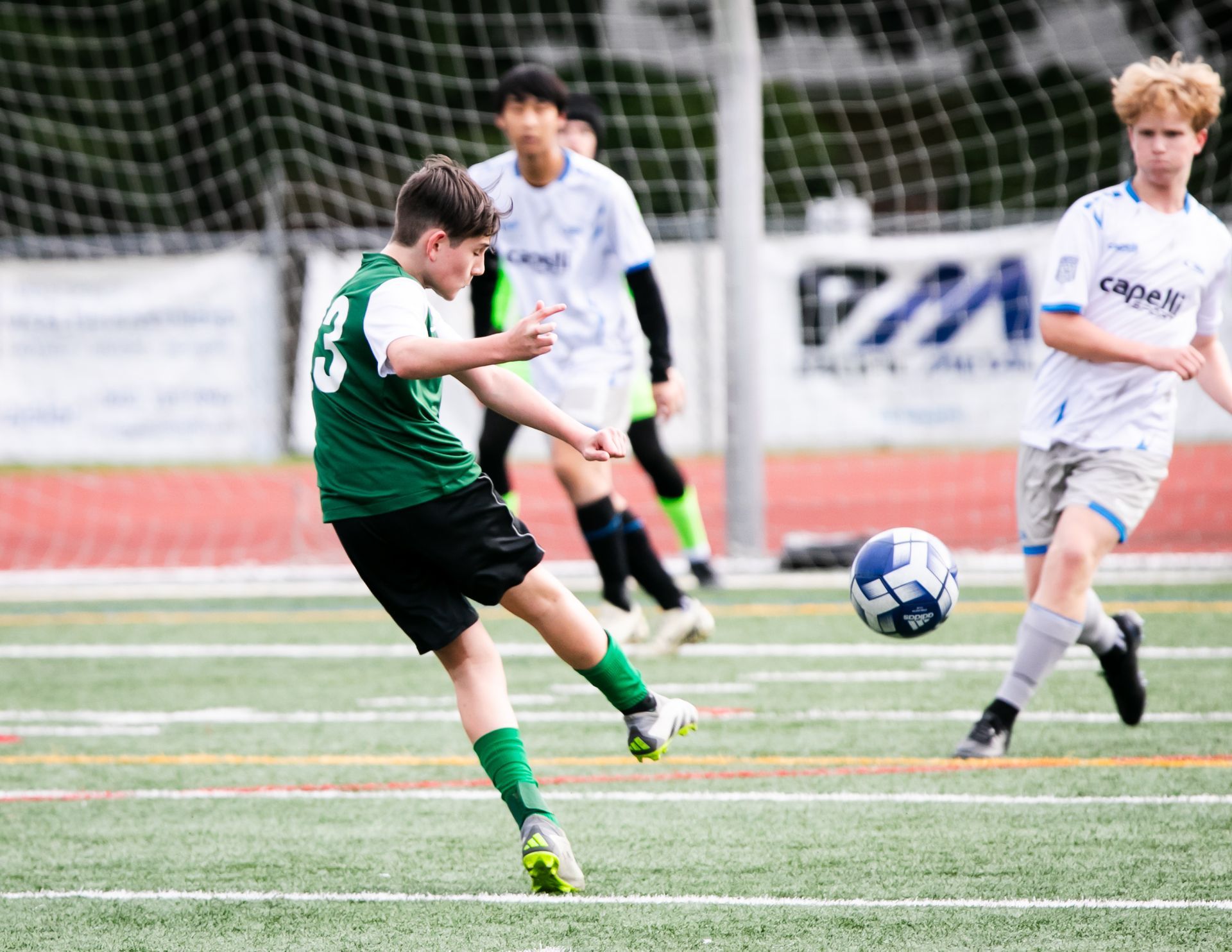 Soccer player in green kicks a ball on a green field, another player in blue watches.