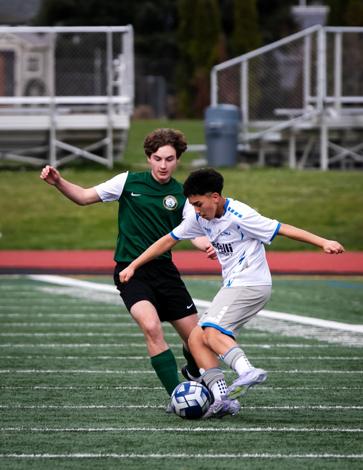 Two soccer players on a green field, one in green jersey, the other in white, both focused on the ball.
