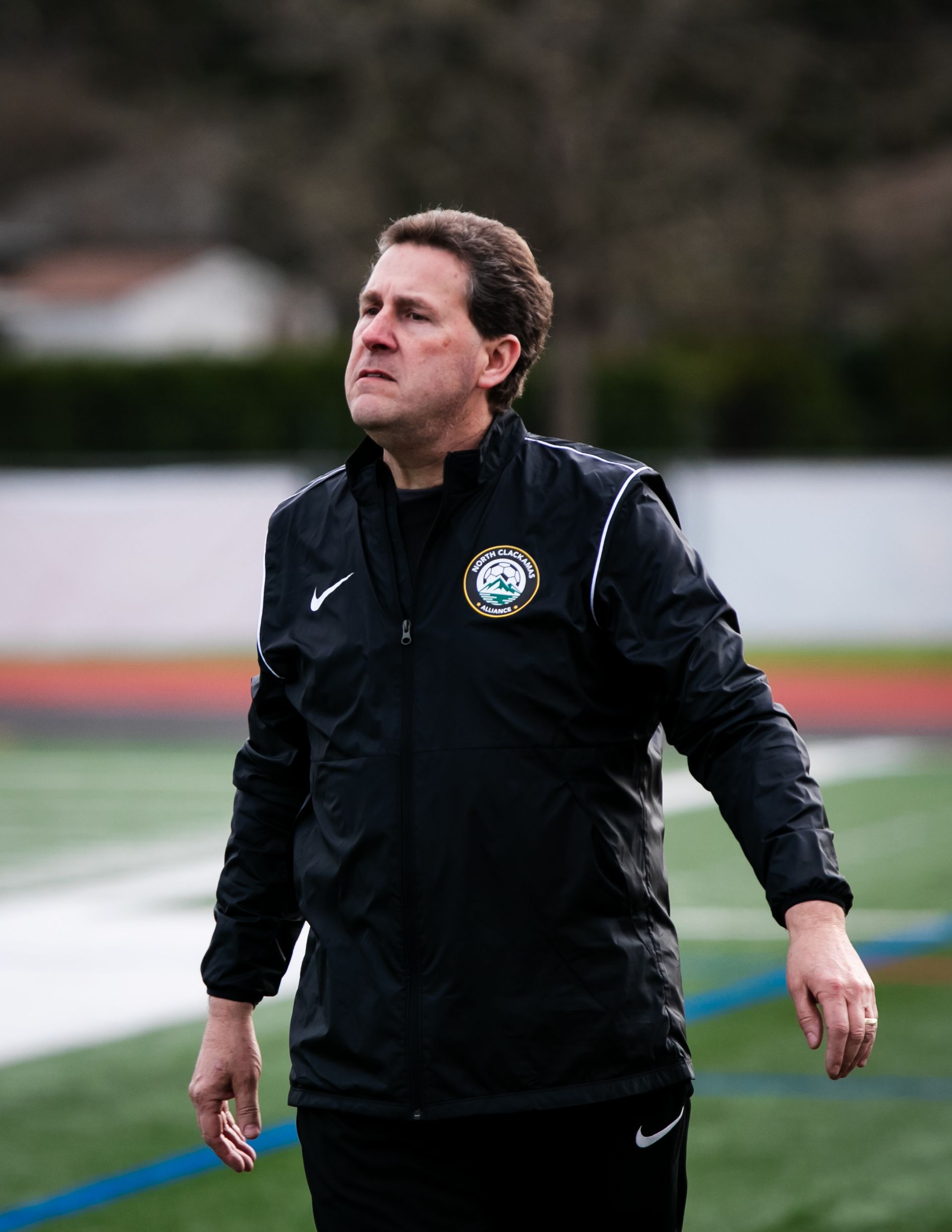 Soccer coach on field wearing a black jacket, looking to the side. Green and white emblem.