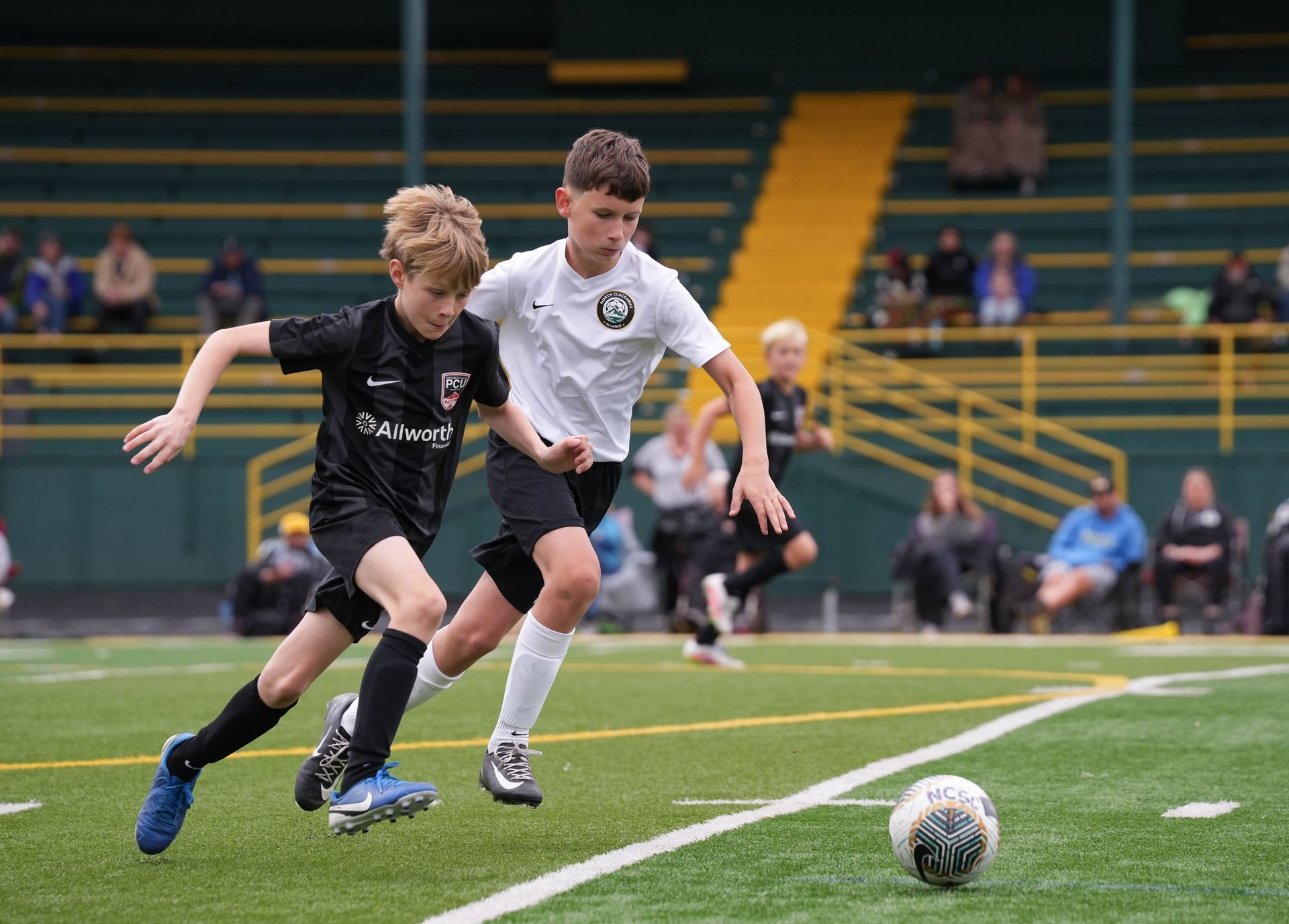 Two boys in soccer uniforms chase the ball on a green field during a game.