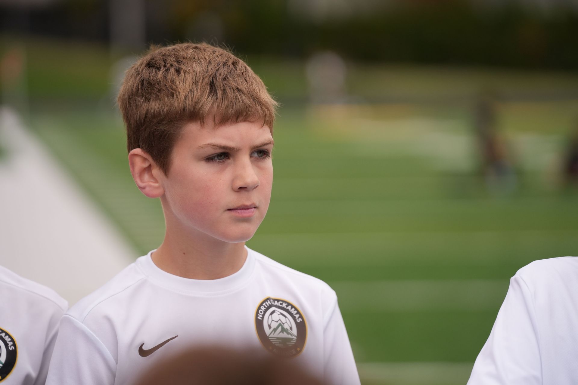 Boy in a white soccer jersey looks toward the left, standing on a green field.