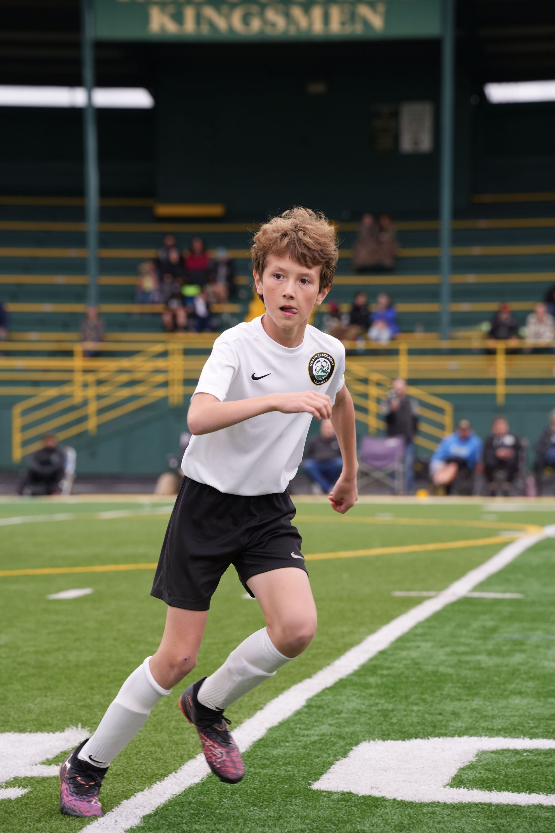 Boy in soccer uniform running on a green field towards the camera. Stands with bleachers in the background.