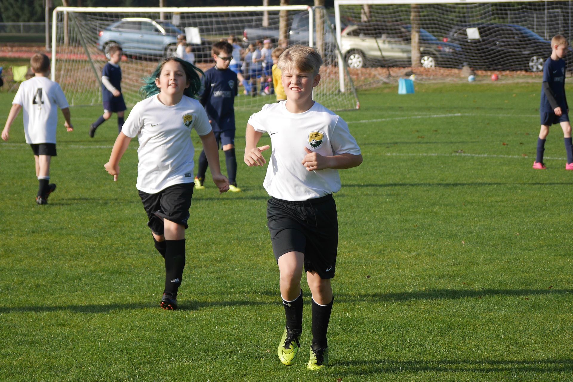 Two young soccer players in white jerseys and black shorts run toward the camera on a green field.