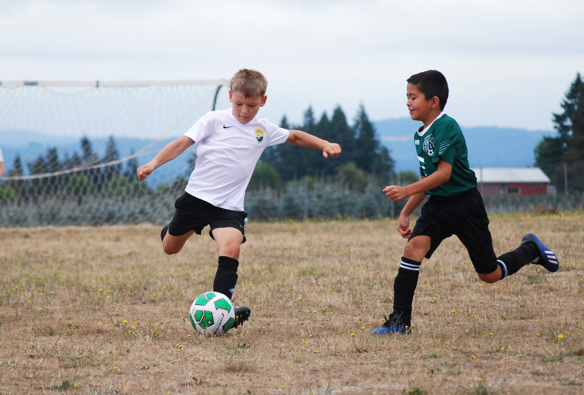 Two boys playing soccer on a dry grass field, one kicking the ball, the other running towards him.