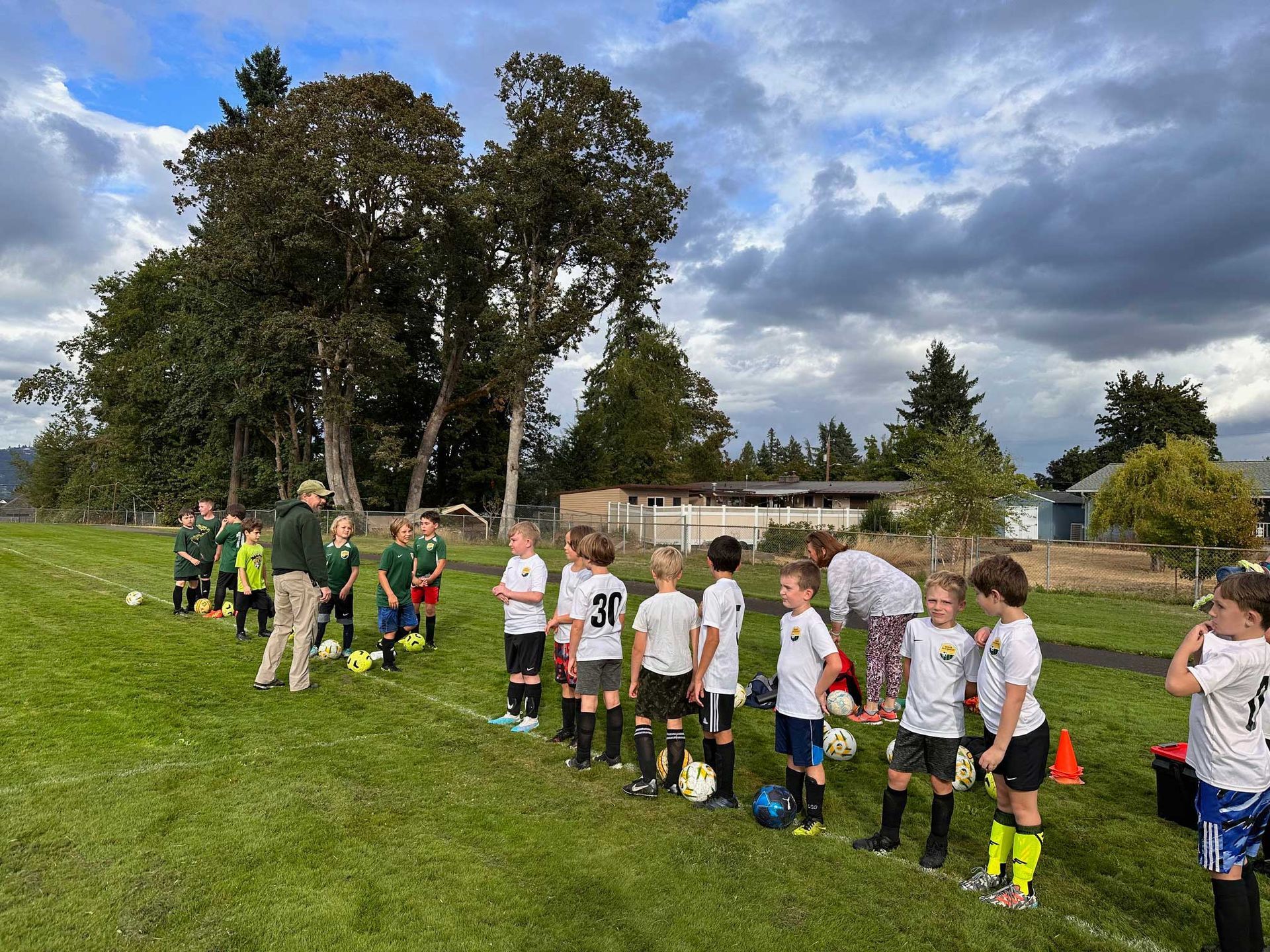 Youth soccer players in white and green jerseys stand on a green field with a coach near trees and a building.