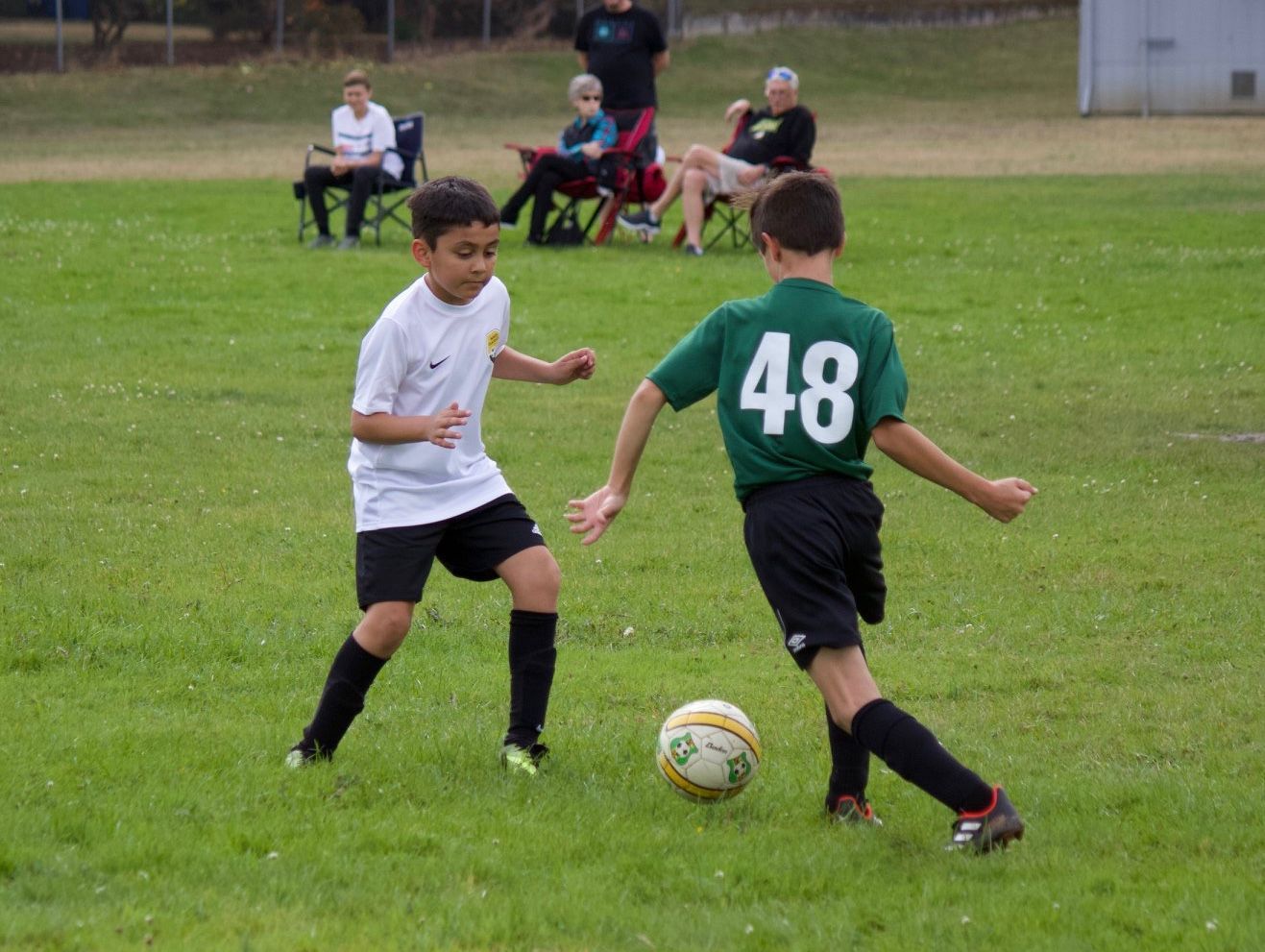 Two boys playing soccer on a green field. One wears a white jersey, the other a green jersey with the number 48. Spectators sit in the background.