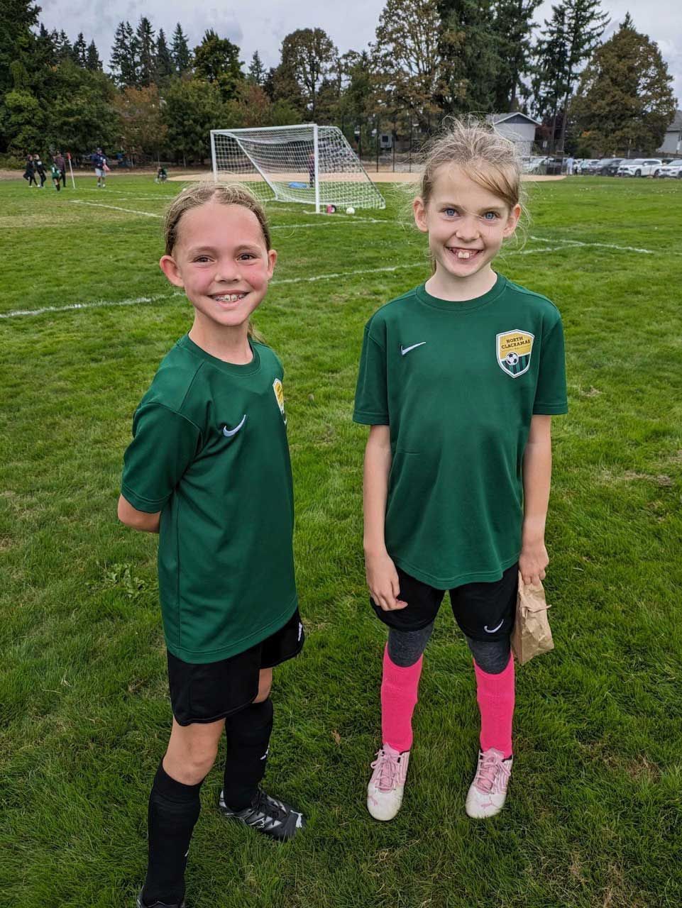 Two young soccer players in green jerseys and black shorts smile on a field. One has pink socks.
