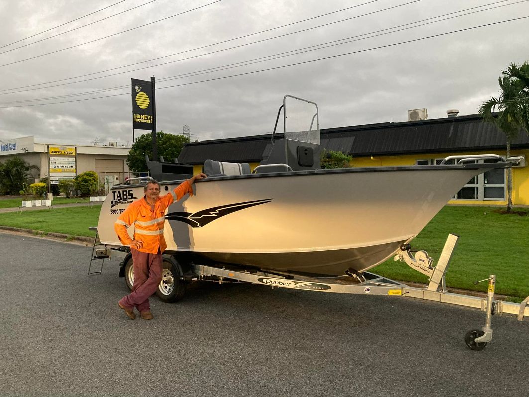 Man In Orange Vest Poses Next To A Silver Boat On A Trailer In Front Of A Building On A Cloudy Day — In Tune Marine in Mackay, QLD