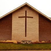 A brick church facade with a stone-textured central panel featuring a large wooden cross above a small stone bench.