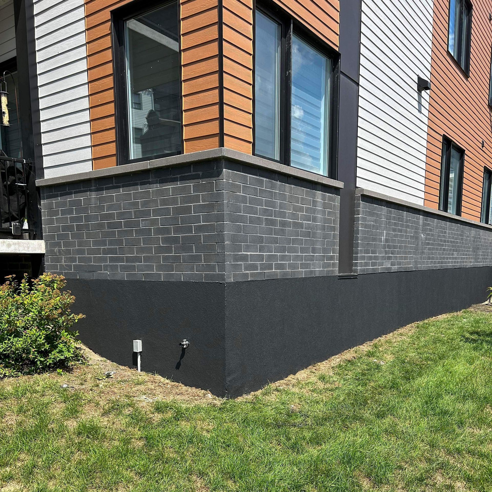 Extérieur d'un bâtiment moderne avec briques sombres et fondations noires. Herbe verte et ciel bleu.