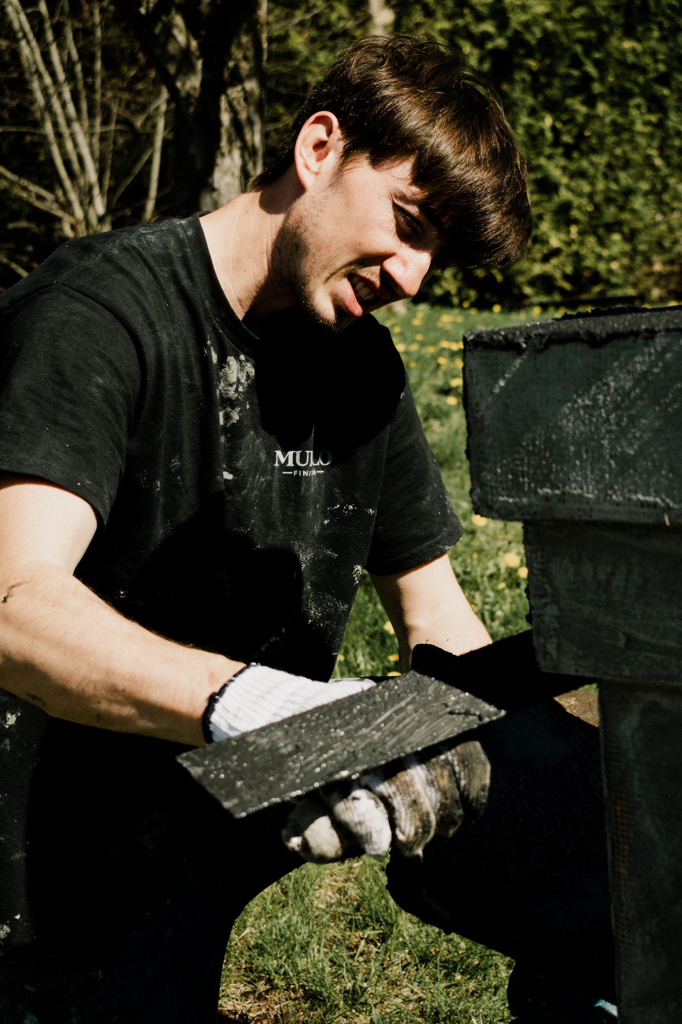 Un homme en chemise noire applique du matériau avec une truelle, travaillant à l'extérieur par une journée ensoleillée.