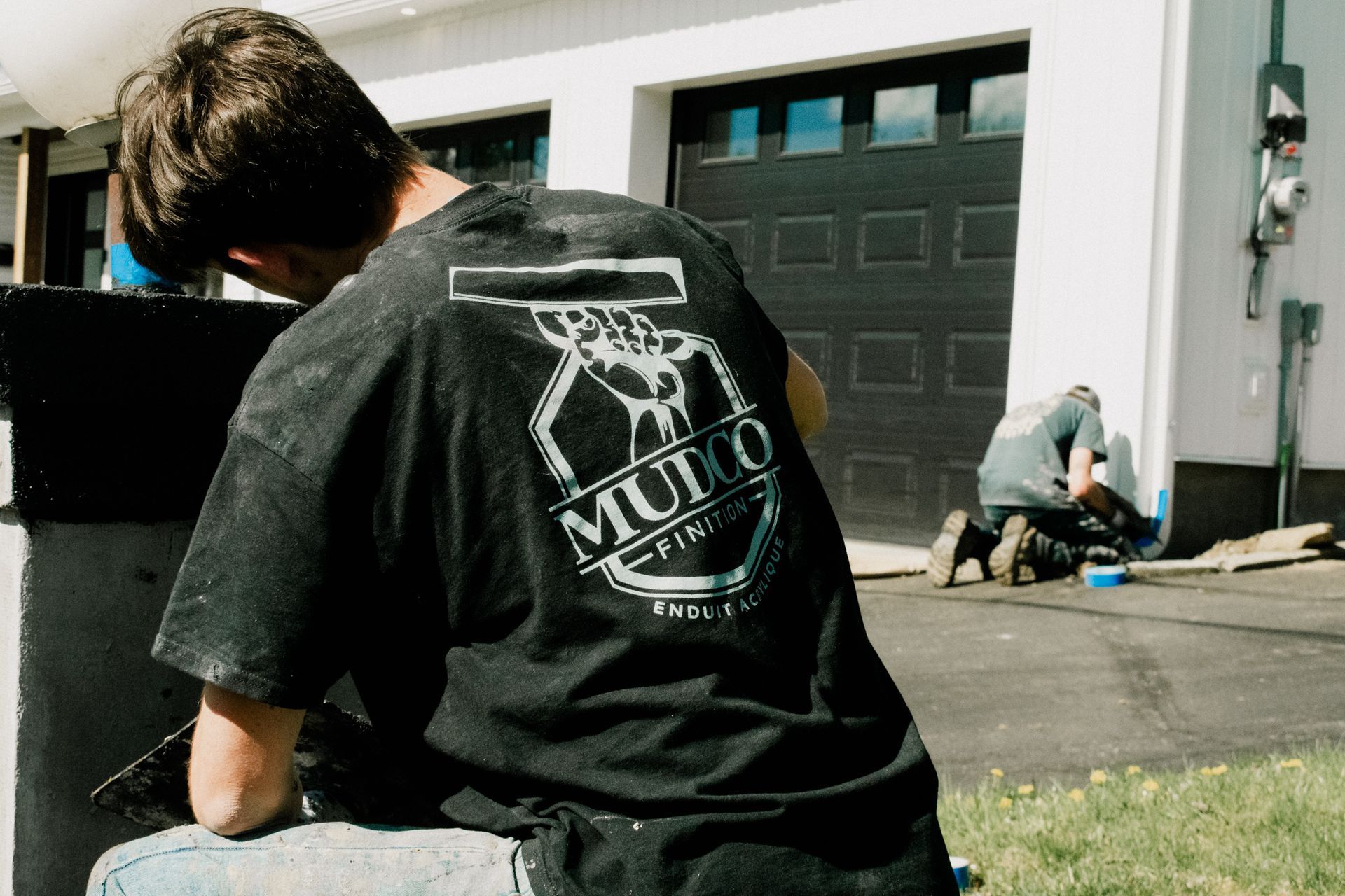 Un homme en chemise noire avec logo travaillant à l'extérieur ; une deuxième personne travaille également en arrière-plan.