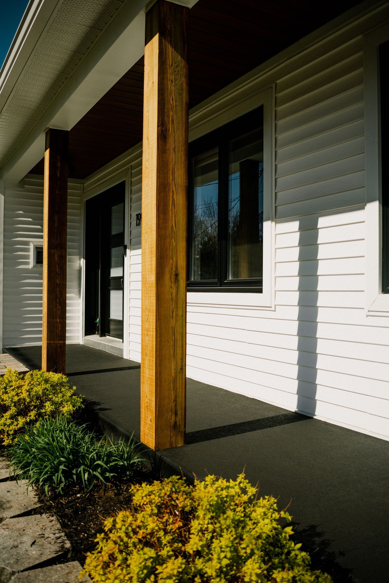 Poteaux de porche en bois sur une maison blanche, avec des buissons et une allée.