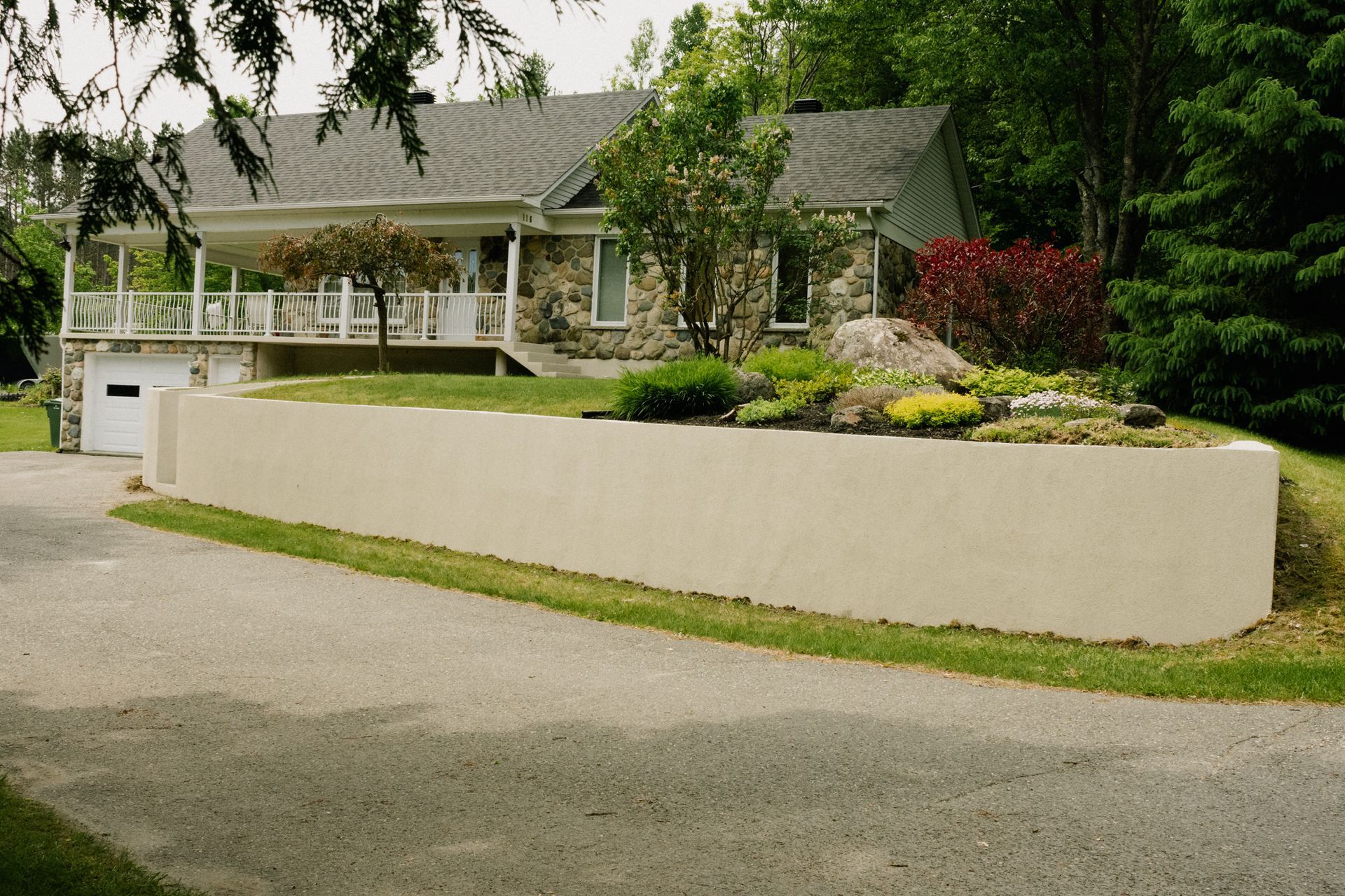 Mur de soutènement beige devant une maison en pierre avec un porche, une porte de garage blanche et une allée.