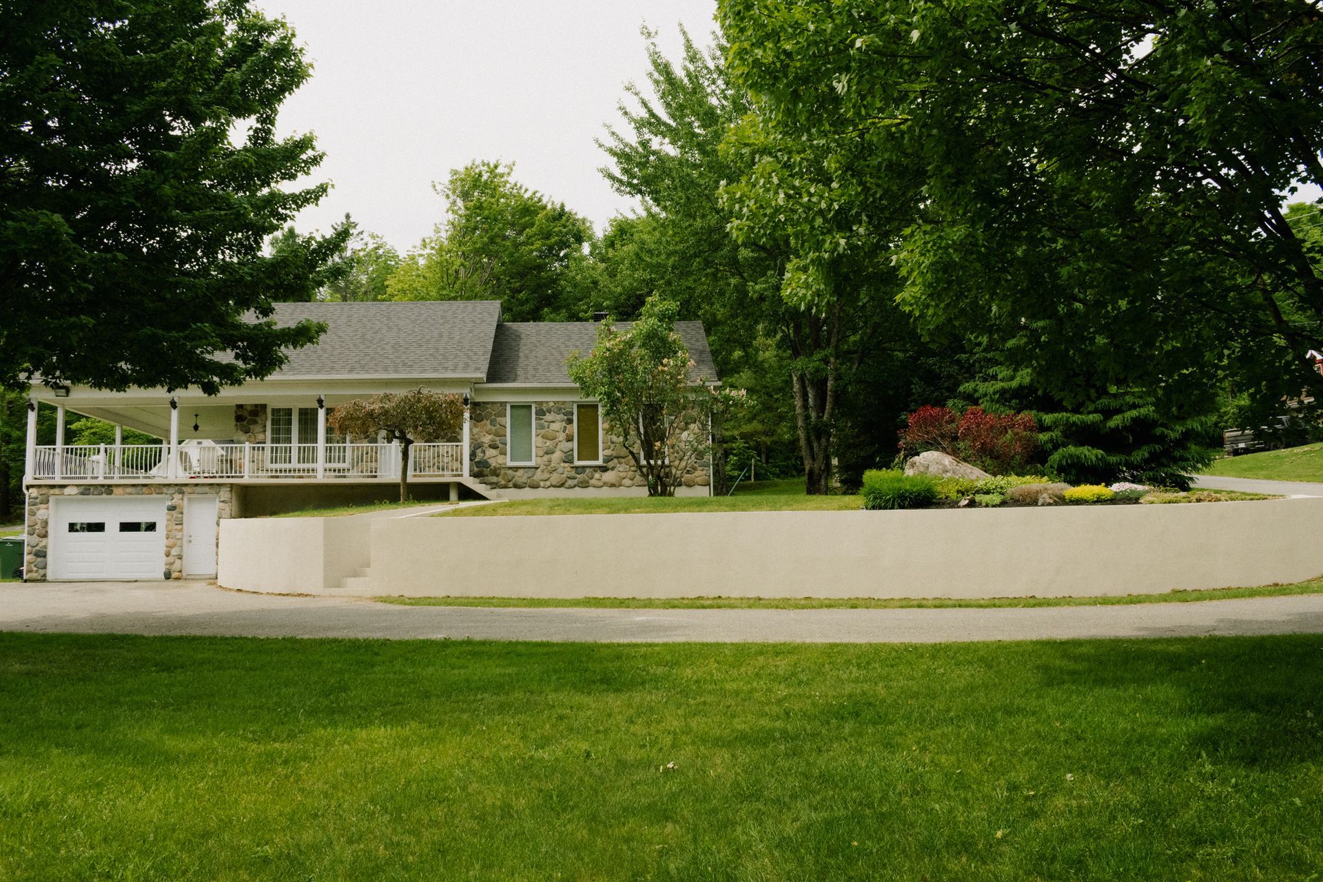 Maison en pierre avec un porche enveloppant et un garage blanc; située dans une pelouse verte avec des arbres.