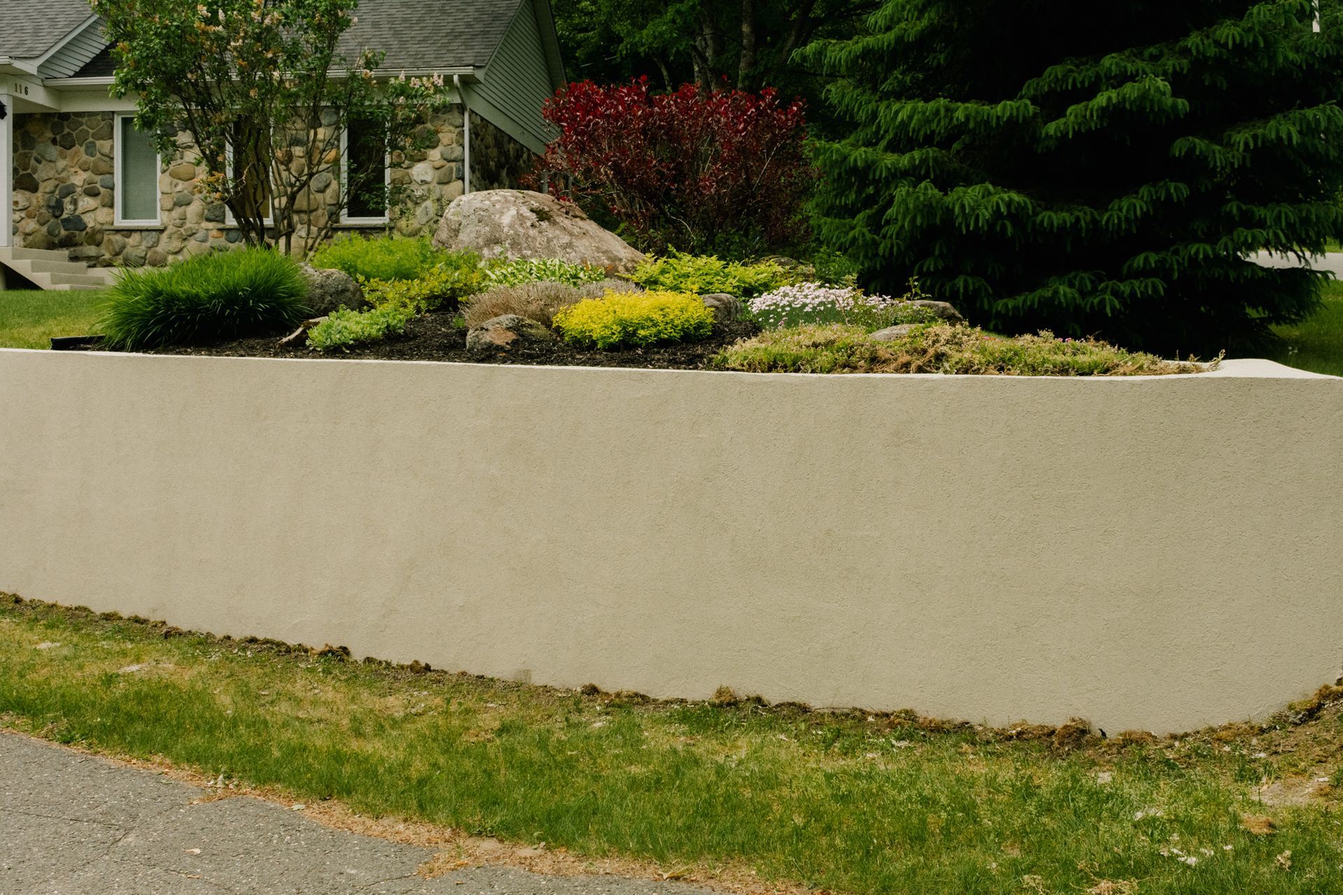 Mur de soutènement beige avec un parterre de plantes luxuriant et une maison en arrière-plan.