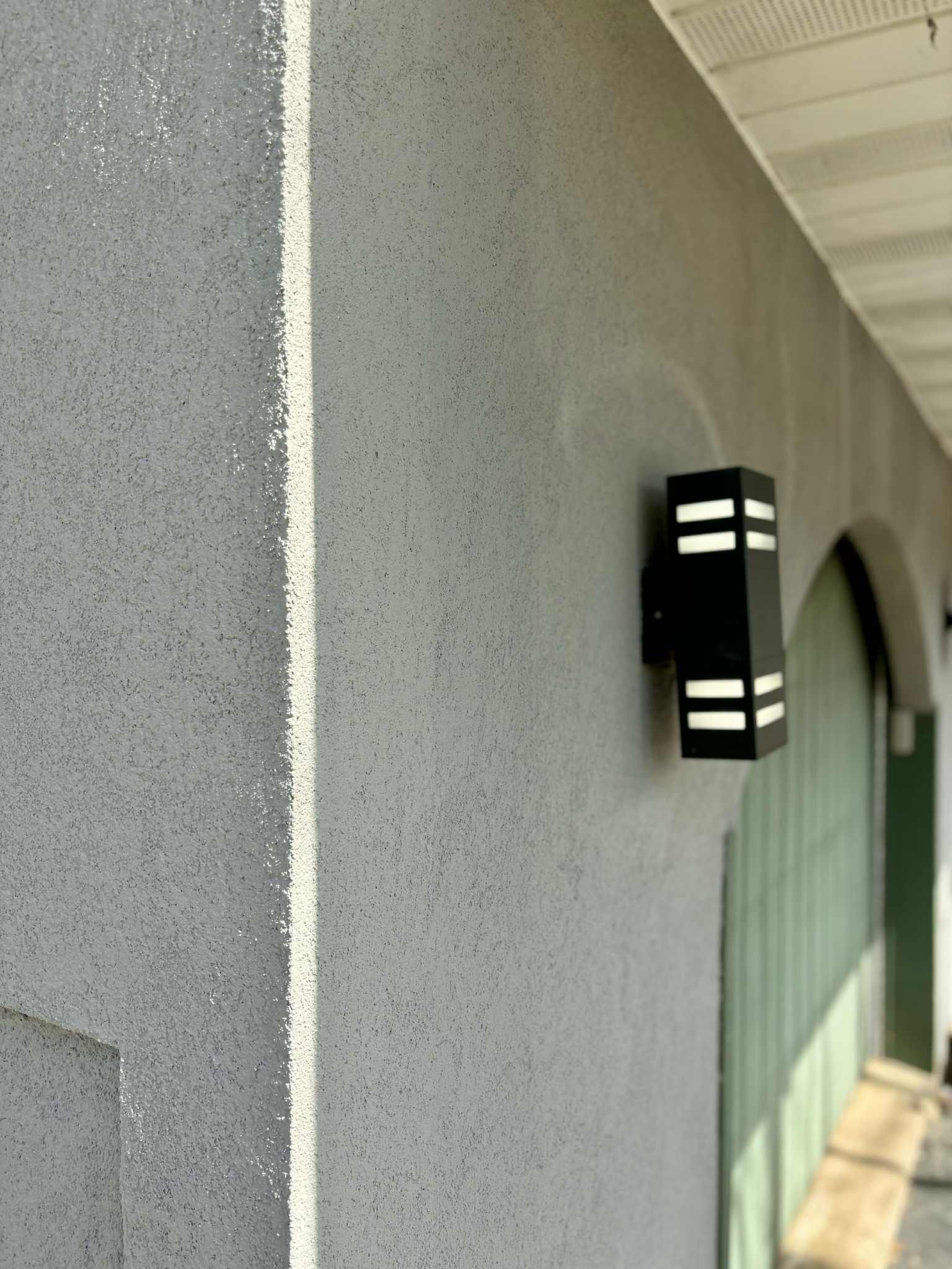 Mur en stuc gris avec luminaire extérieur noir et fenêtre verte visible.