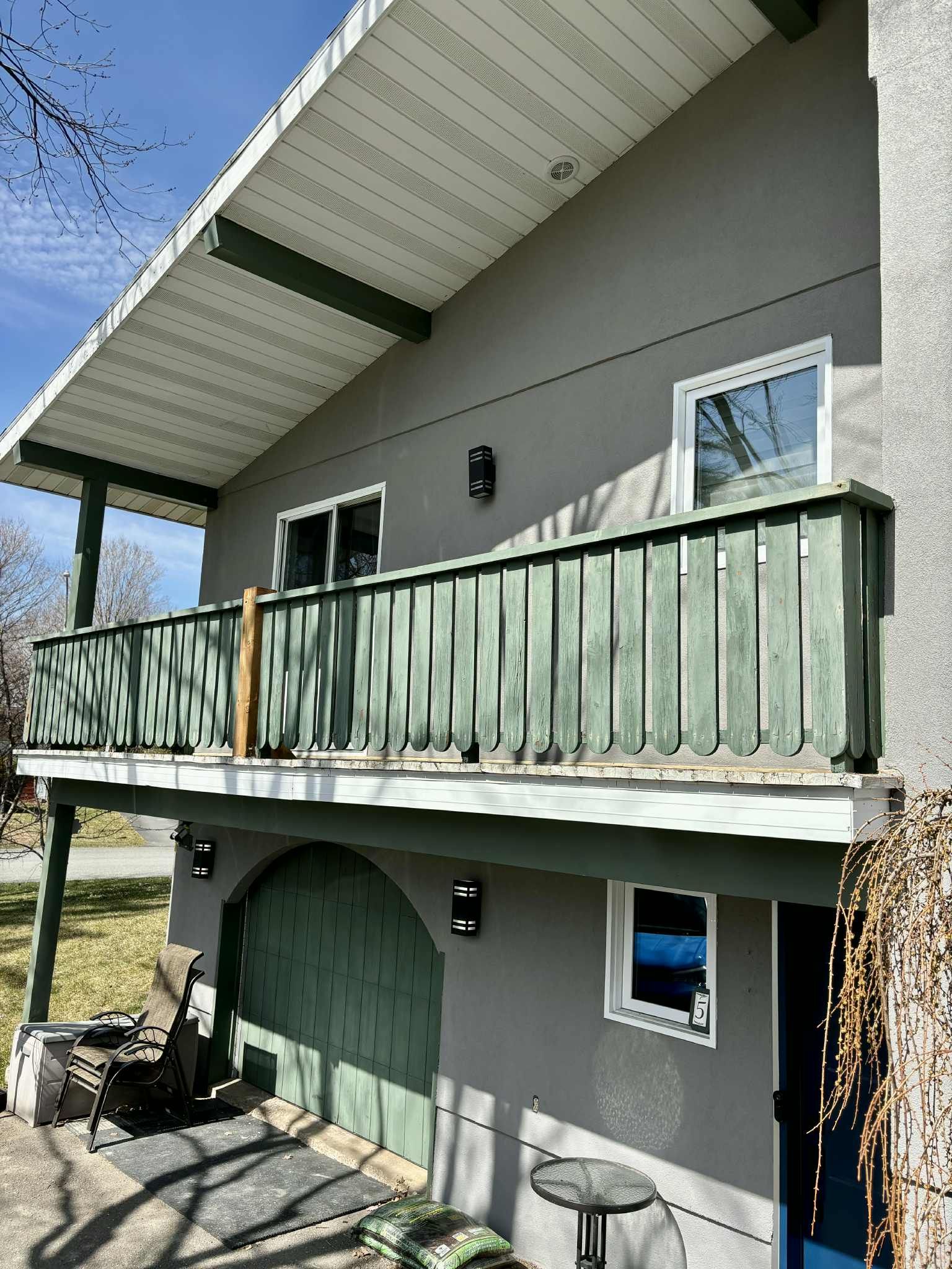 Maison grise avec balcon vert, porte cintrée et garniture blanche sous un ciel bleu.