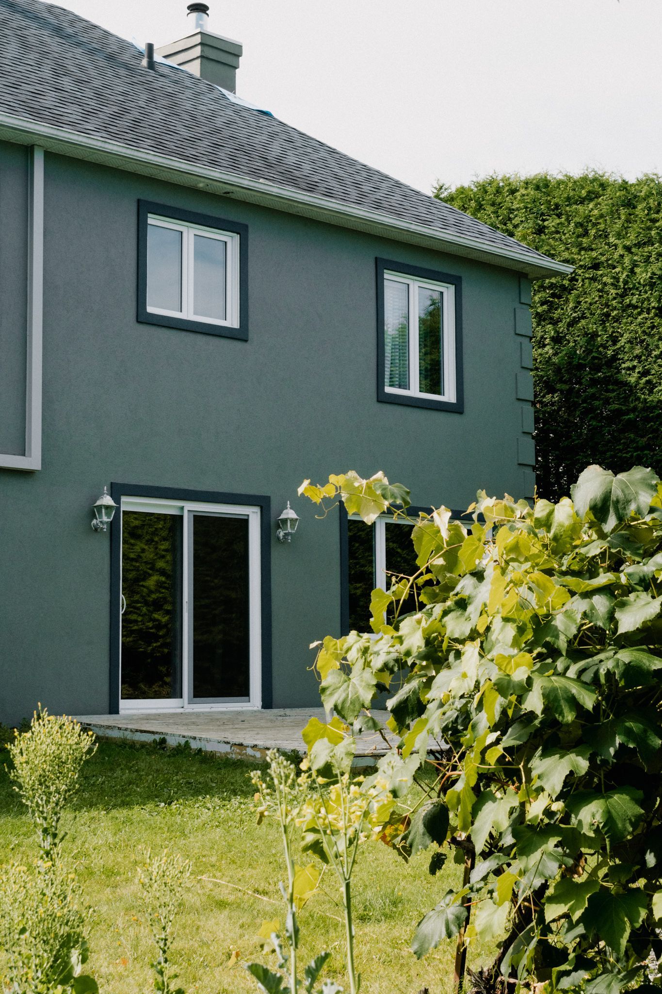 Maison à deux étages avec revêtement gris et fenêtres à cadre blanc. Une porte-fenêtre coulissante donne sur un jardin verdoyant.