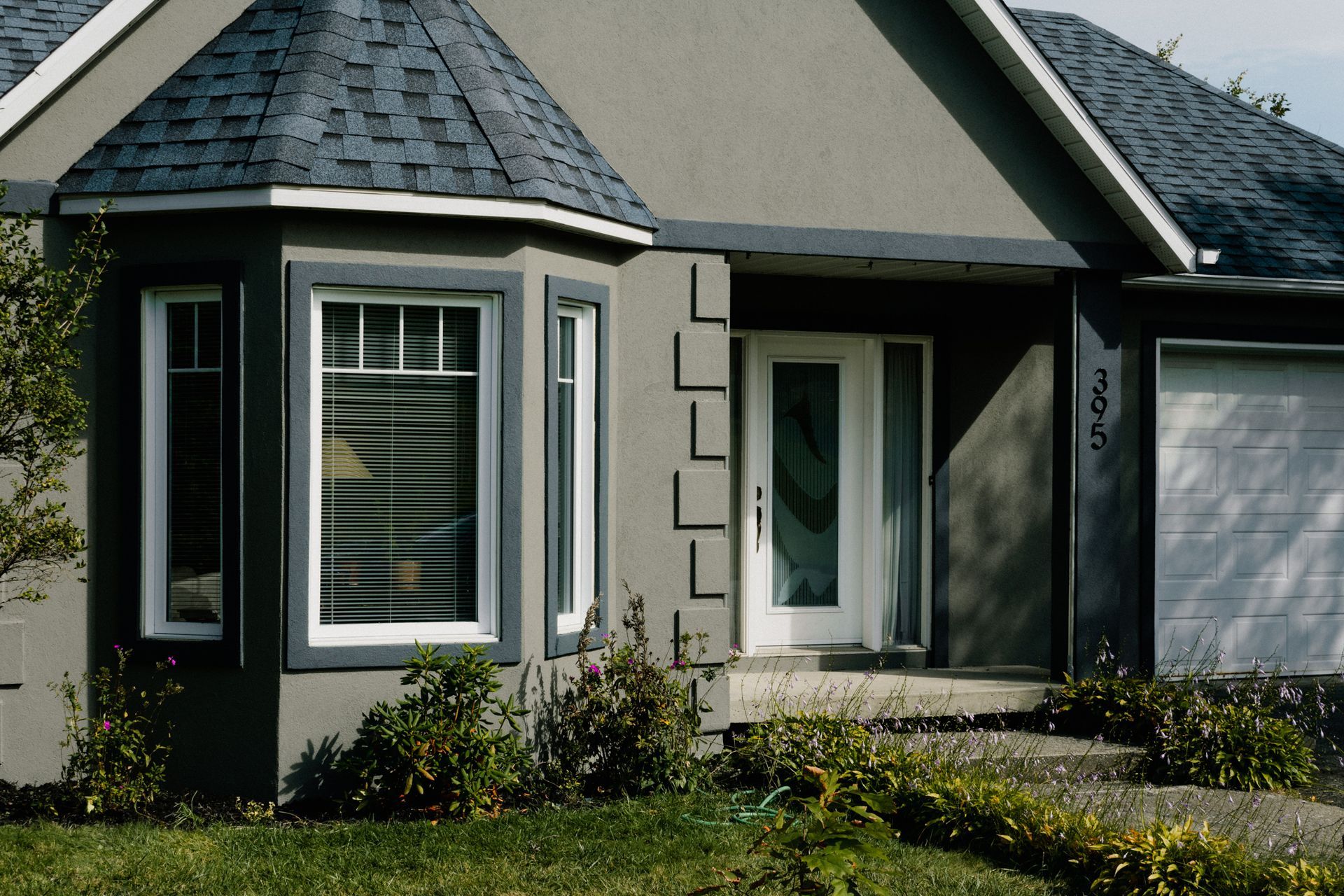 Extérieur de la maison avec baie vitrée, porte d'entrée et garage ; palette de couleurs gris et blanc.