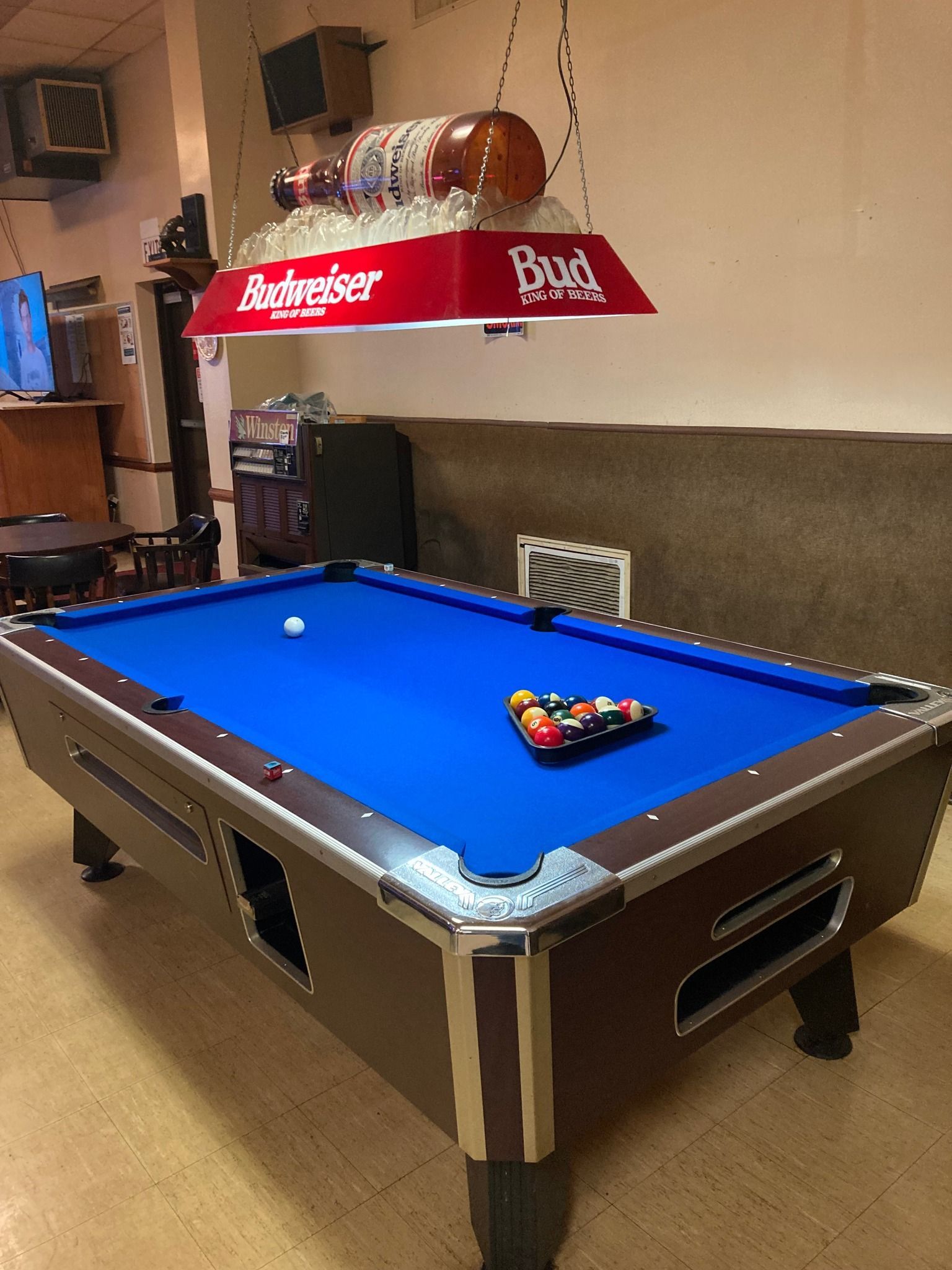 A blue pool table with a racked set of balls stands under a red Budweiser hanging lamp in a room with tan flooring.